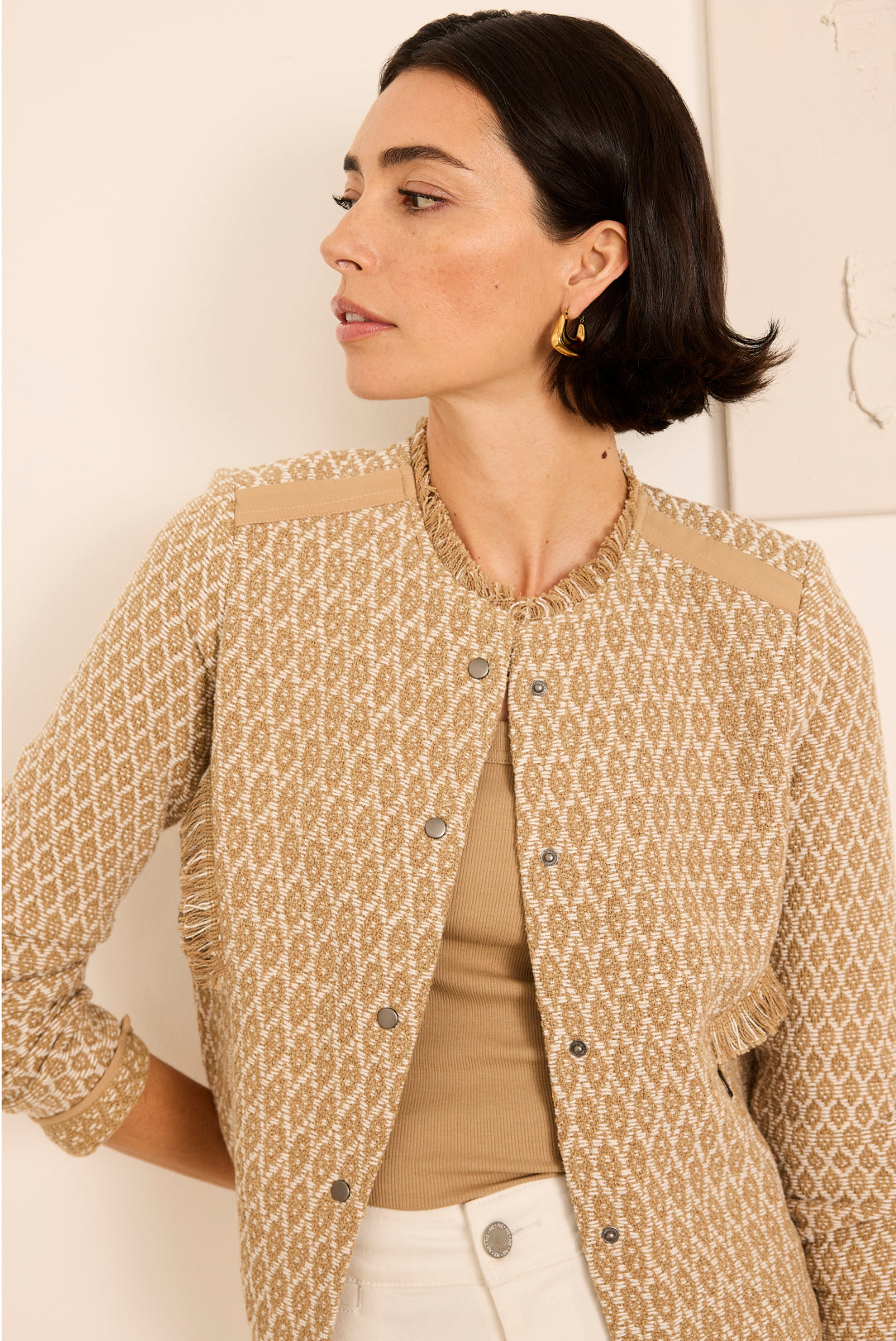 Woman wearing a patterned beige jacket over a brown top and white pants, standing against a light background.