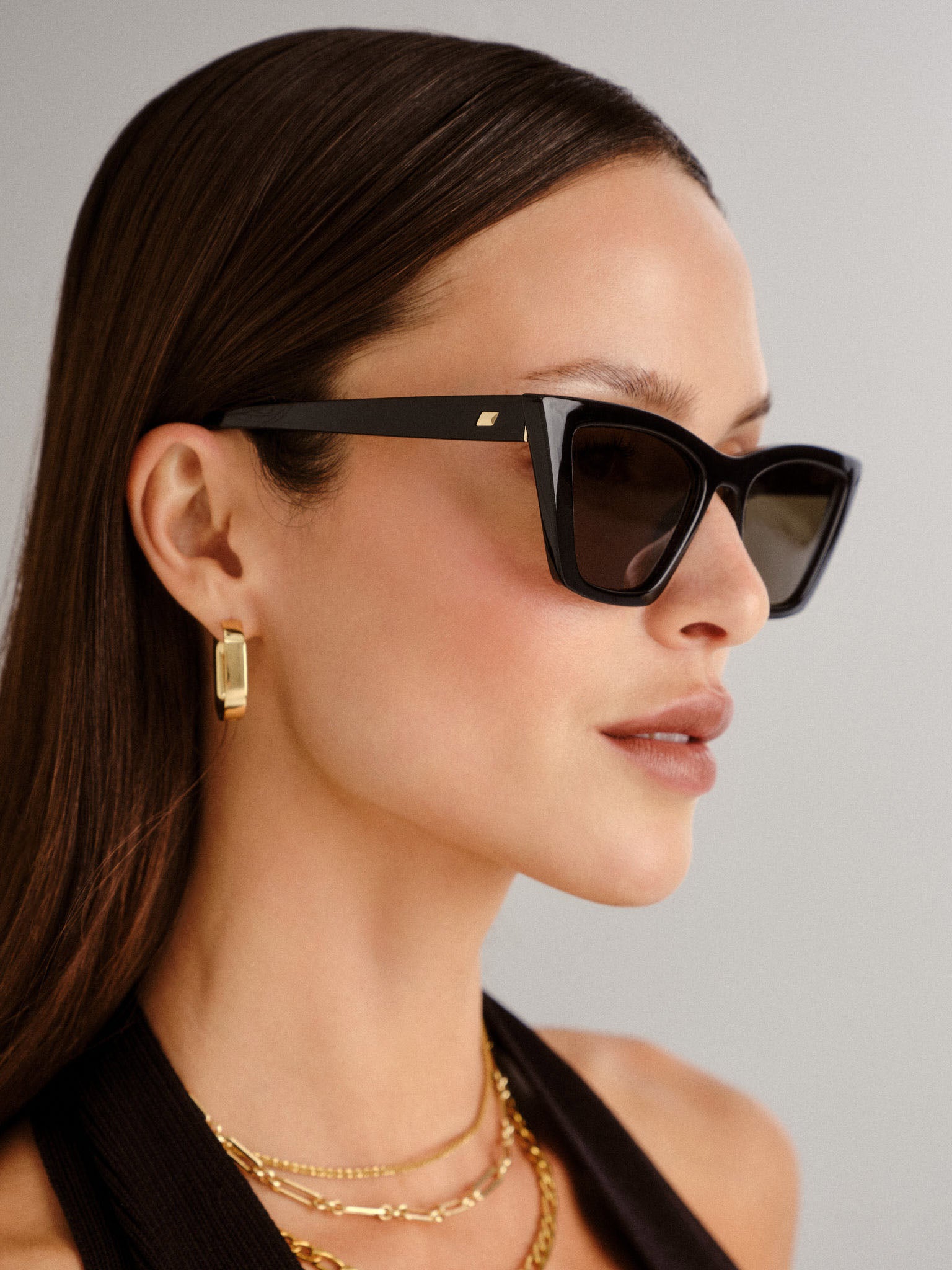 A model wears bold black rectangular sunglasses with thick frames, dark lenses, and a subtle gold logo at the temple.