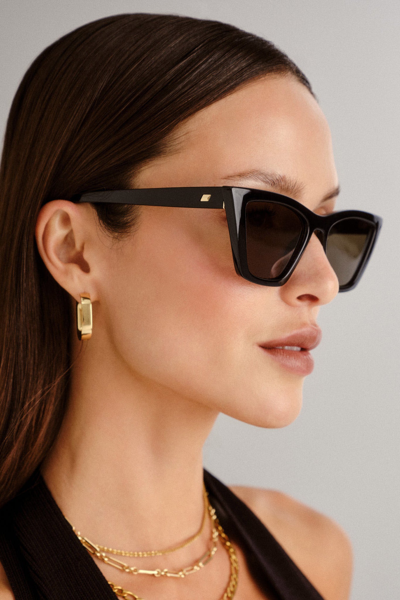 A model wears bold black rectangular sunglasses with thick frames, dark lenses, and a subtle gold logo at the temple.