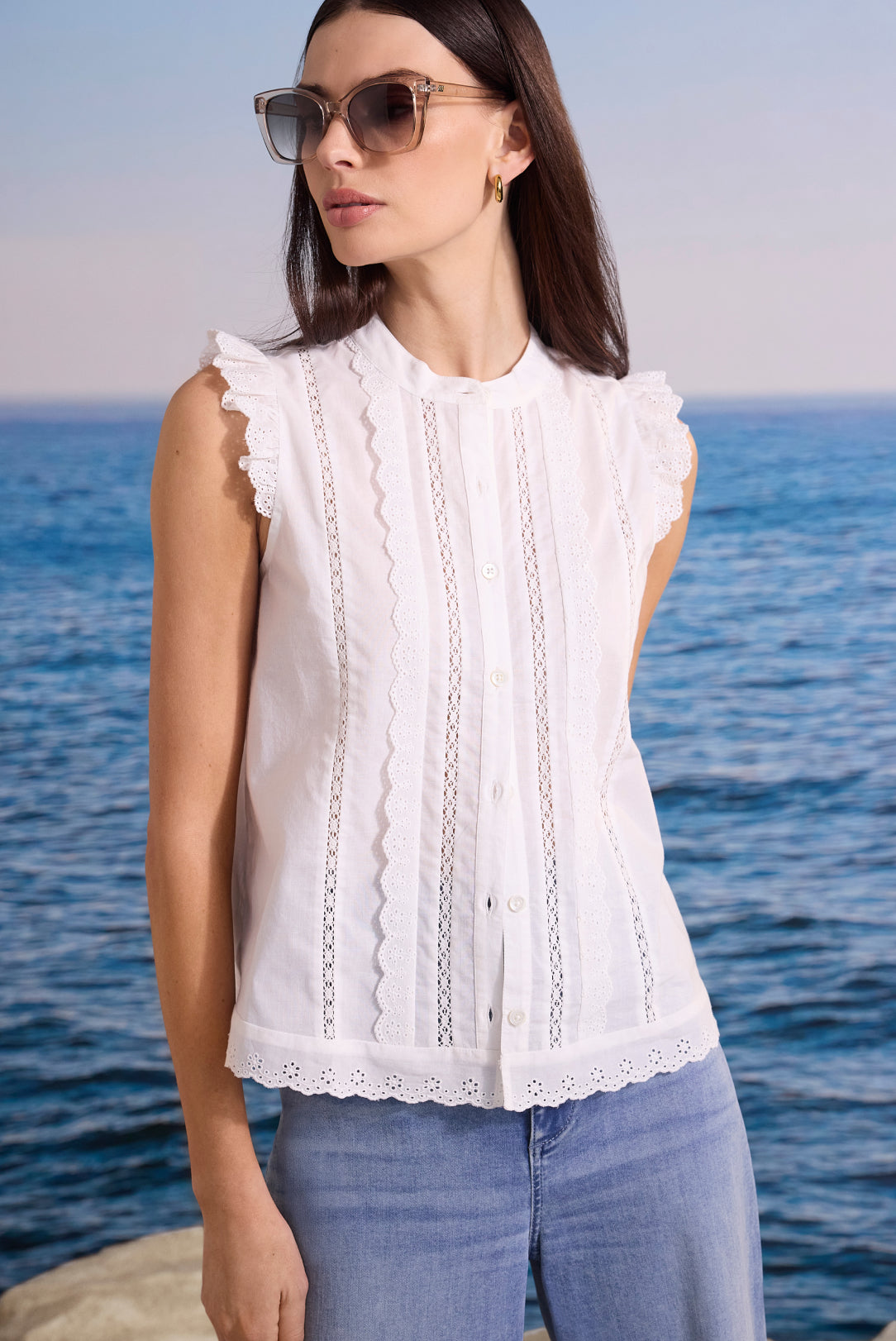 Woman wearing a white sleeveless top with ruffled details and sunglasses, standing by the ocean.