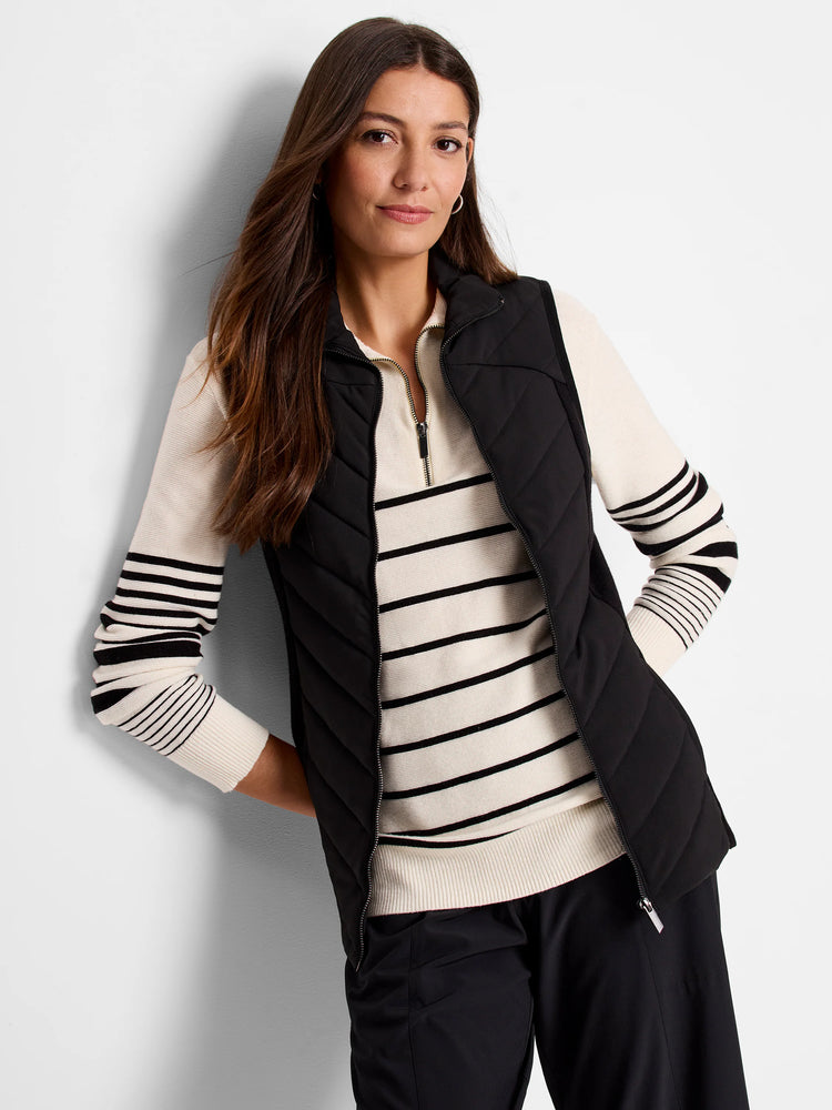 Black quilted puffer vest with zipper. Diagonal quilting pattern. Model wears it over a cream sweater with black stripes.