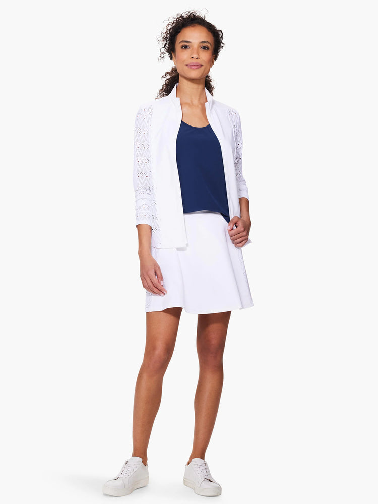 White seamed tank and jacket with eyelet detail; navy blue undershirt; white skirt. Model is wearing white sneakers.