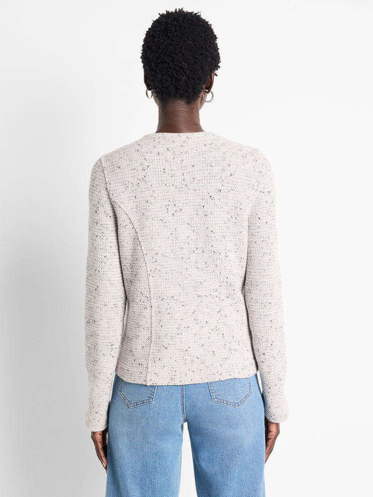 A fitted, collarless cream boucle jacket featuring dark flecks, long sleeves, and shaping seams is seen from the back.