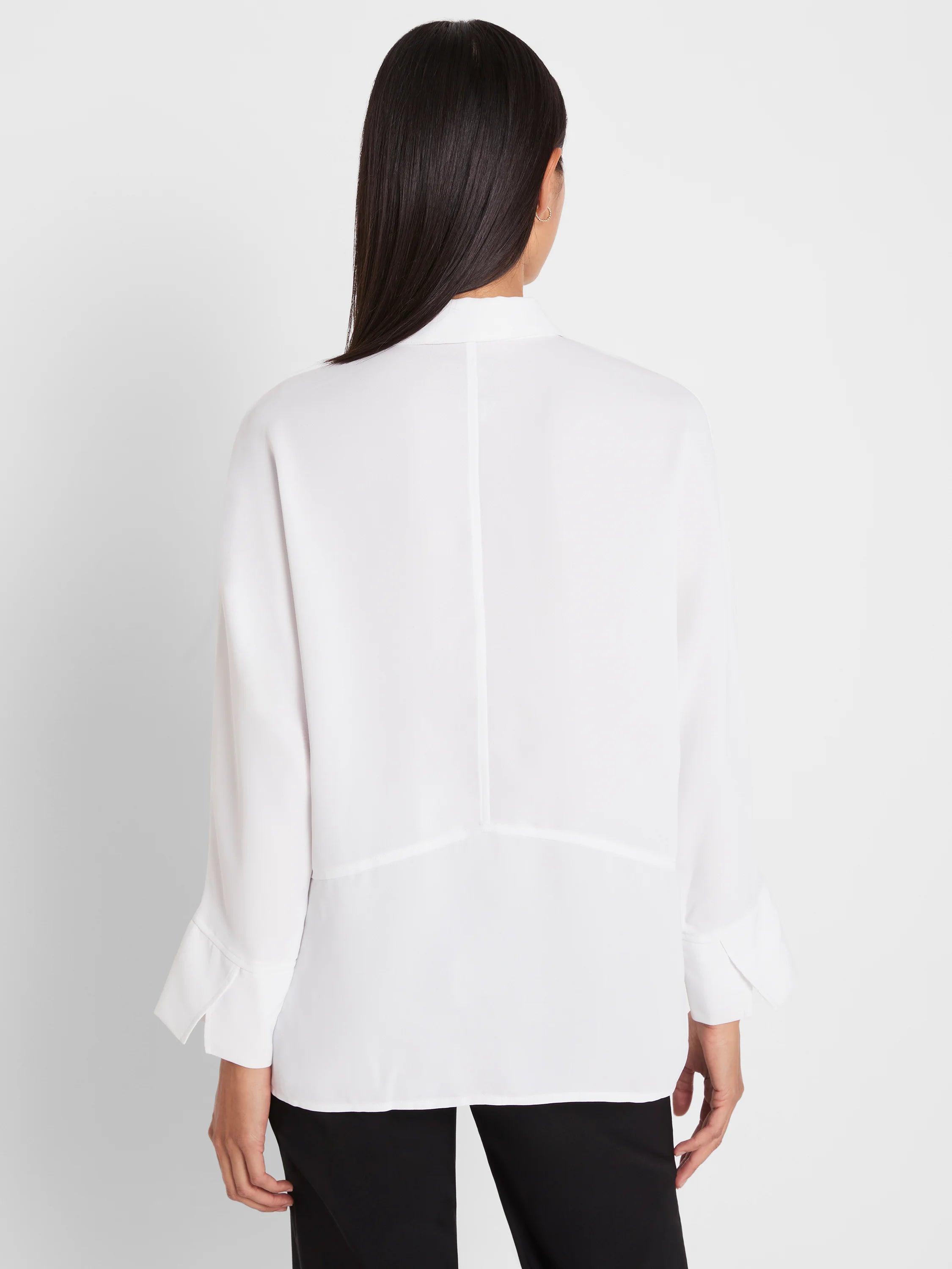 White loose-fitting blouse, back view. Features vertical back seam, horizontal seam detail, and wide cuffs.