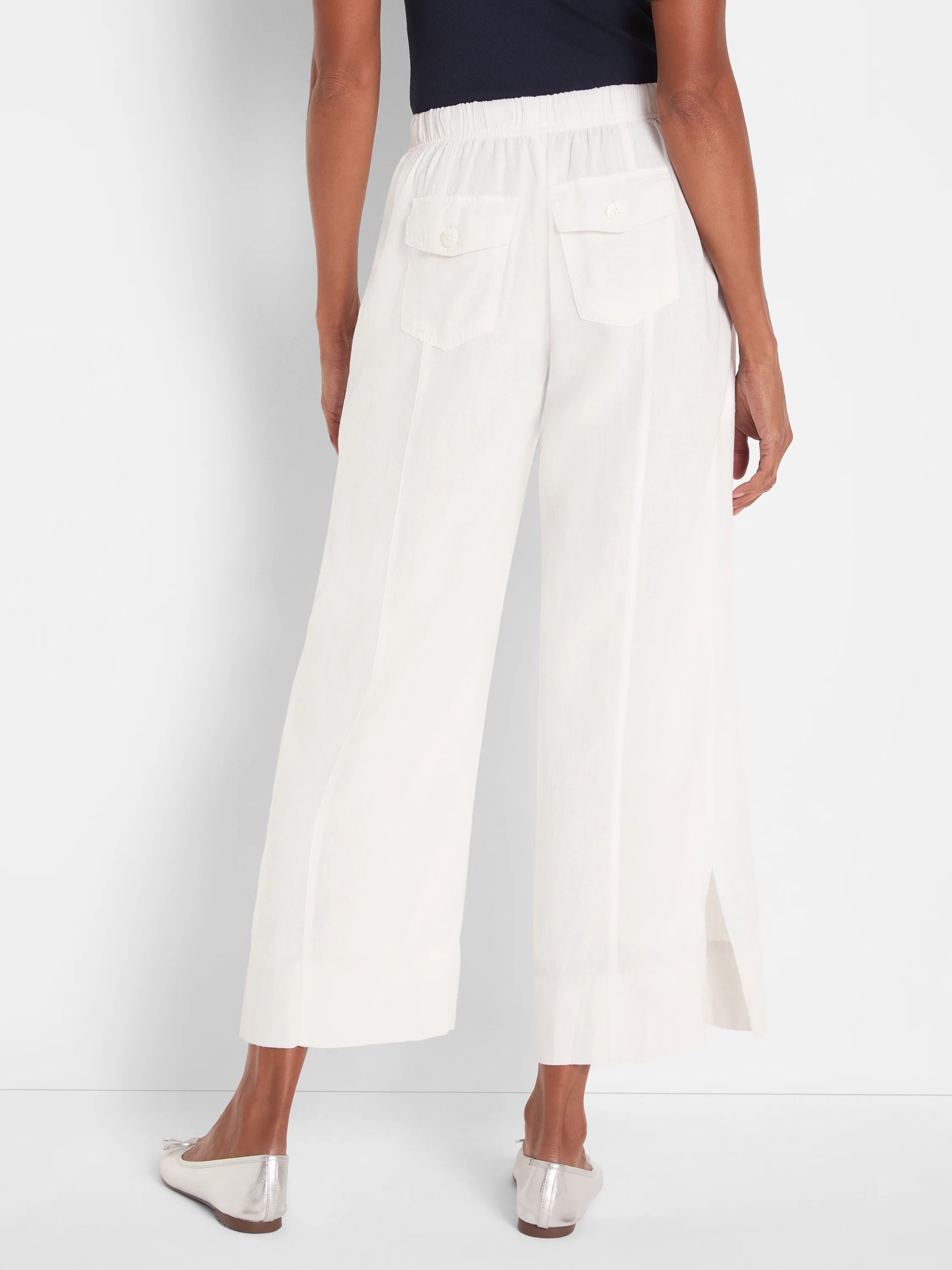 White wide-leg crop pants, rear view. Elastic back waist, two buttoned flap pockets, vertical leg seams, and side hem slits.