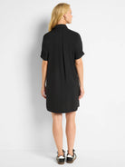 Black shirt dress, back view. Collared, short cuffed sleeves, loose-fitting, above-knee length, with back pleats.
