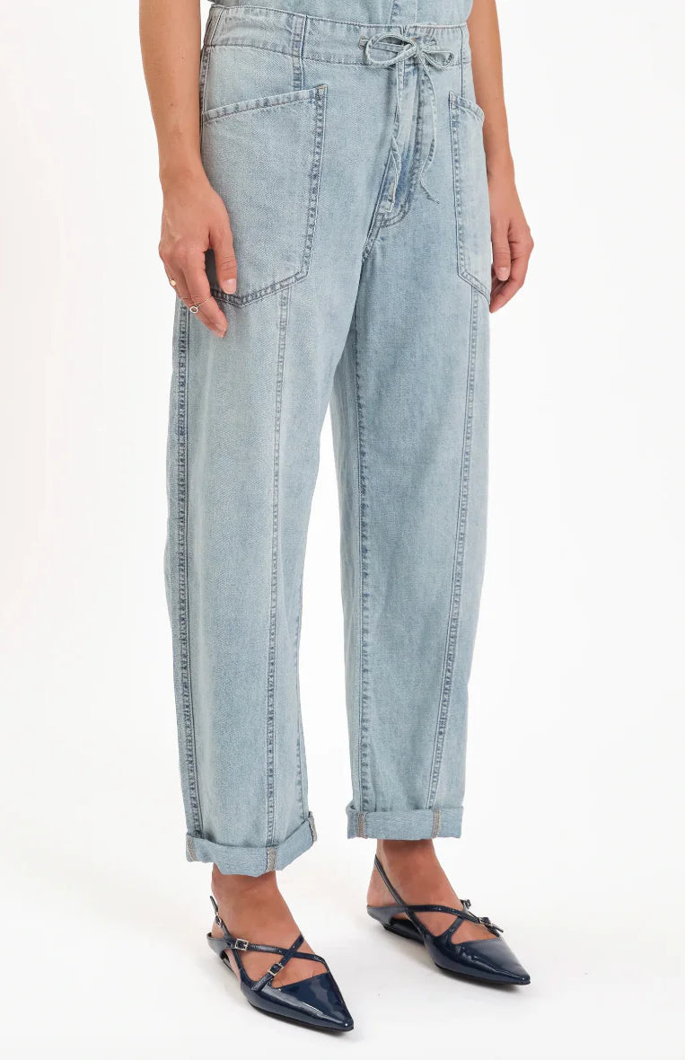 Light wash blue barrel leg jeans with tied drawstring, large front patch pockets, exposed vertical seams, shown cuffed.