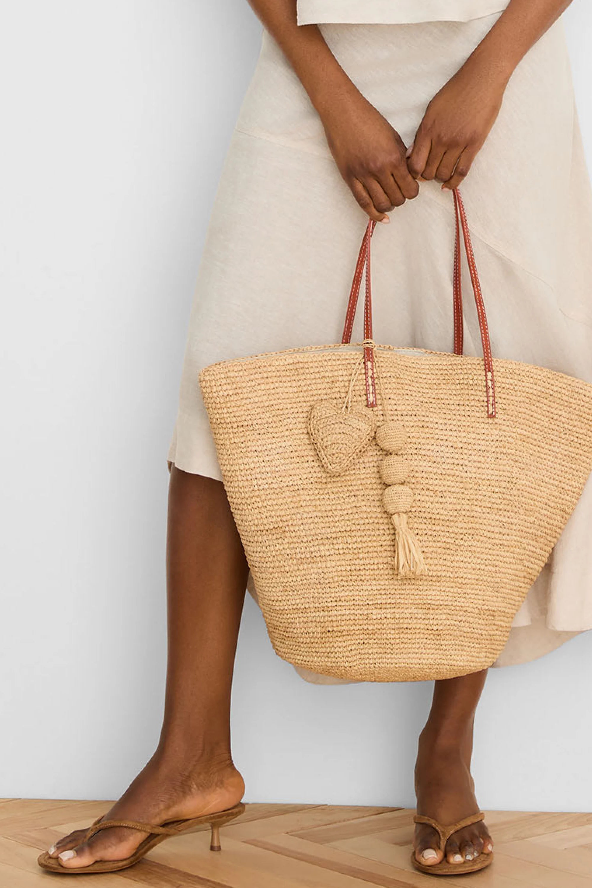 Beige woven tote bag with dark brown straps. Features a matching woven heart charm & a beaded tassel charm.