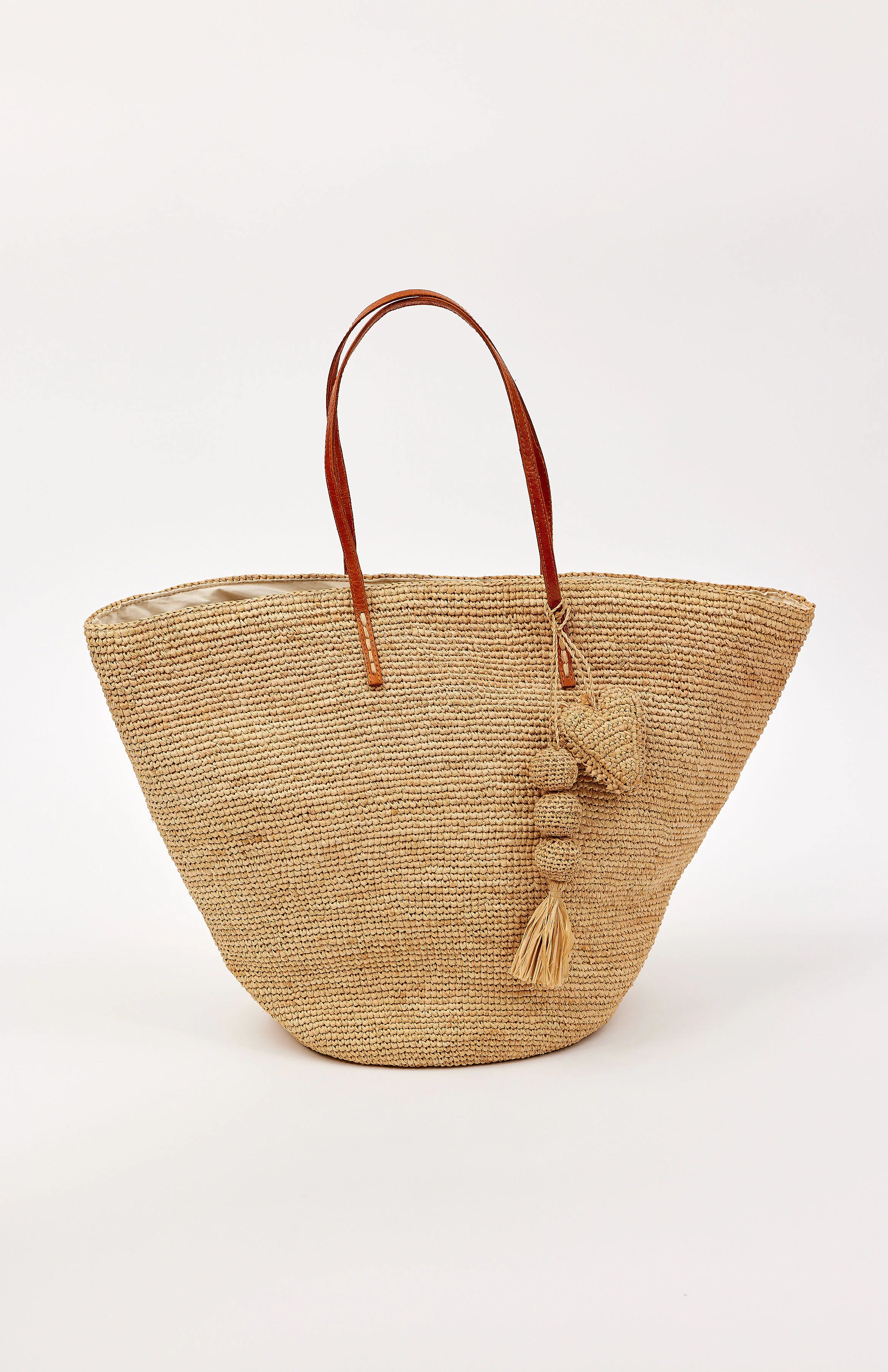Tapered natural straw tote bag with brown leather handles. Features straw balls, tassel, and heart charms.