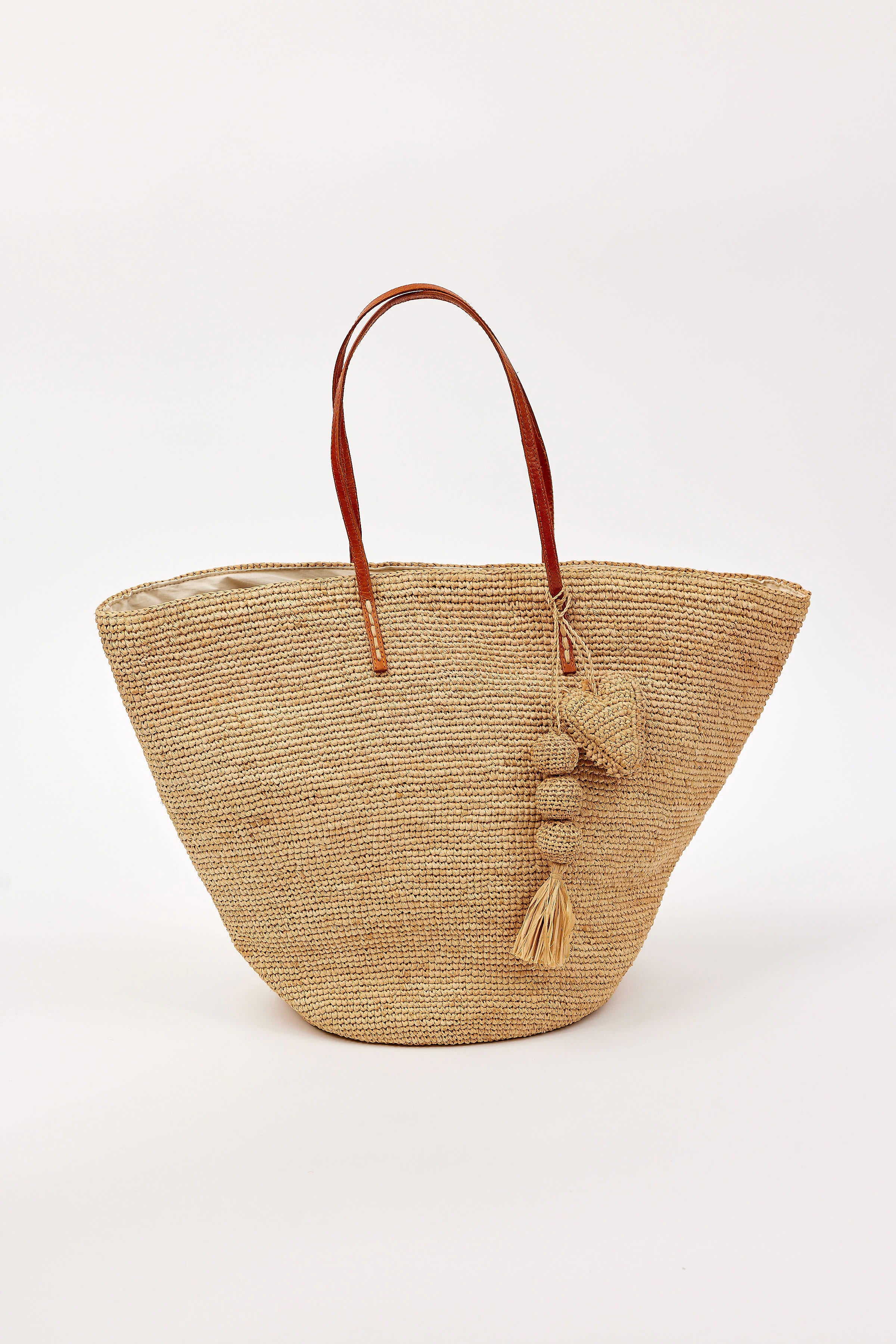 Tapered natural straw tote bag with brown leather handles. Features straw balls, tassel, and heart charms.