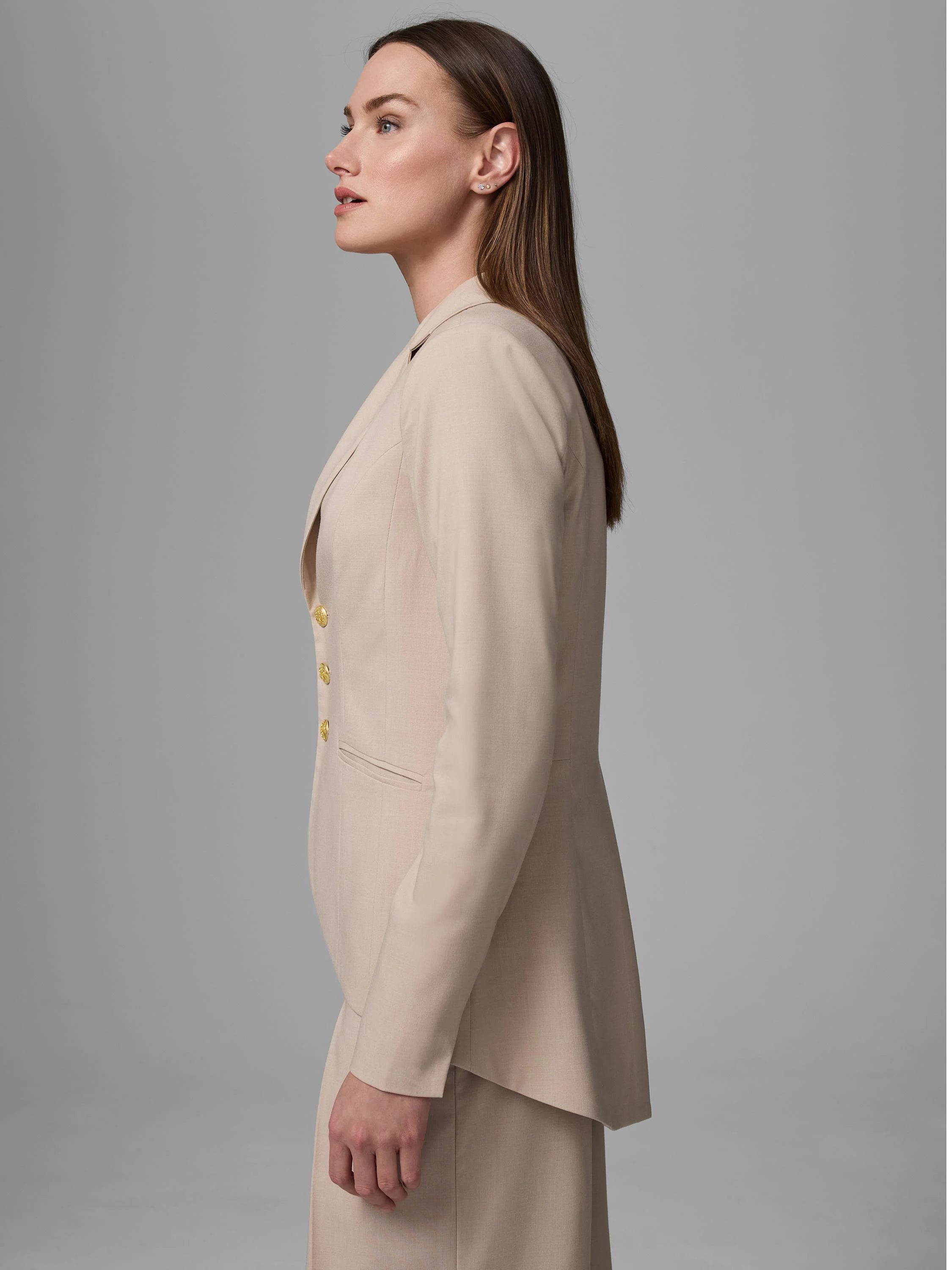 Light beige blazer with gold buttons, notched lapel, and flap pockets, worn by a woman in side profile.