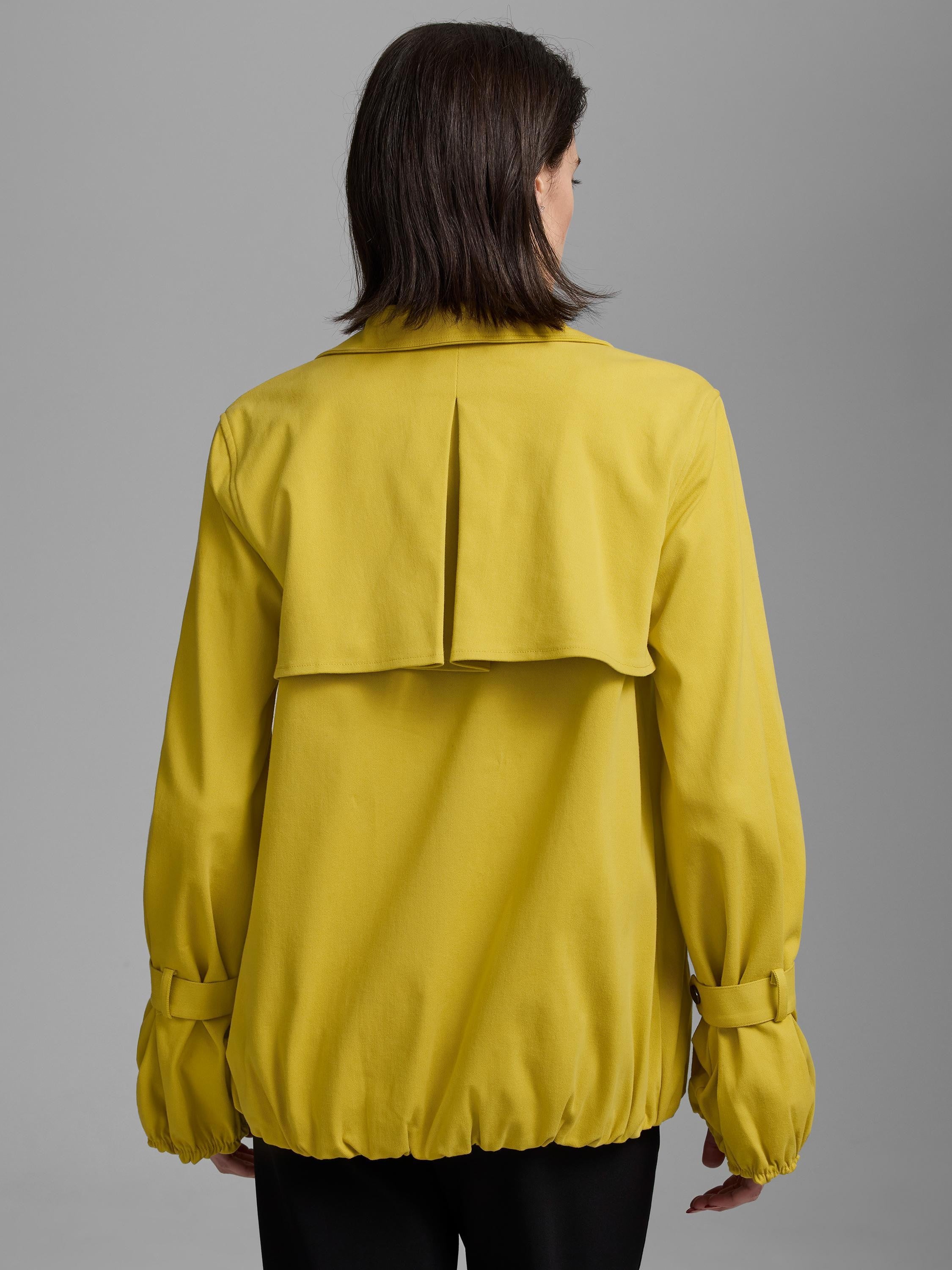Back of a woman wearing a yellow twill trench coat with a pleated storm flap, buckled cuffs, and an elasticated hem.