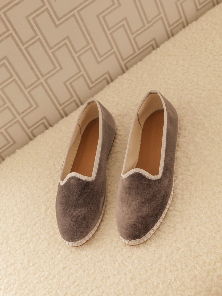 Gray velvet slippers with off-white trim and beige interior, shown on an off-white fluffy surface.