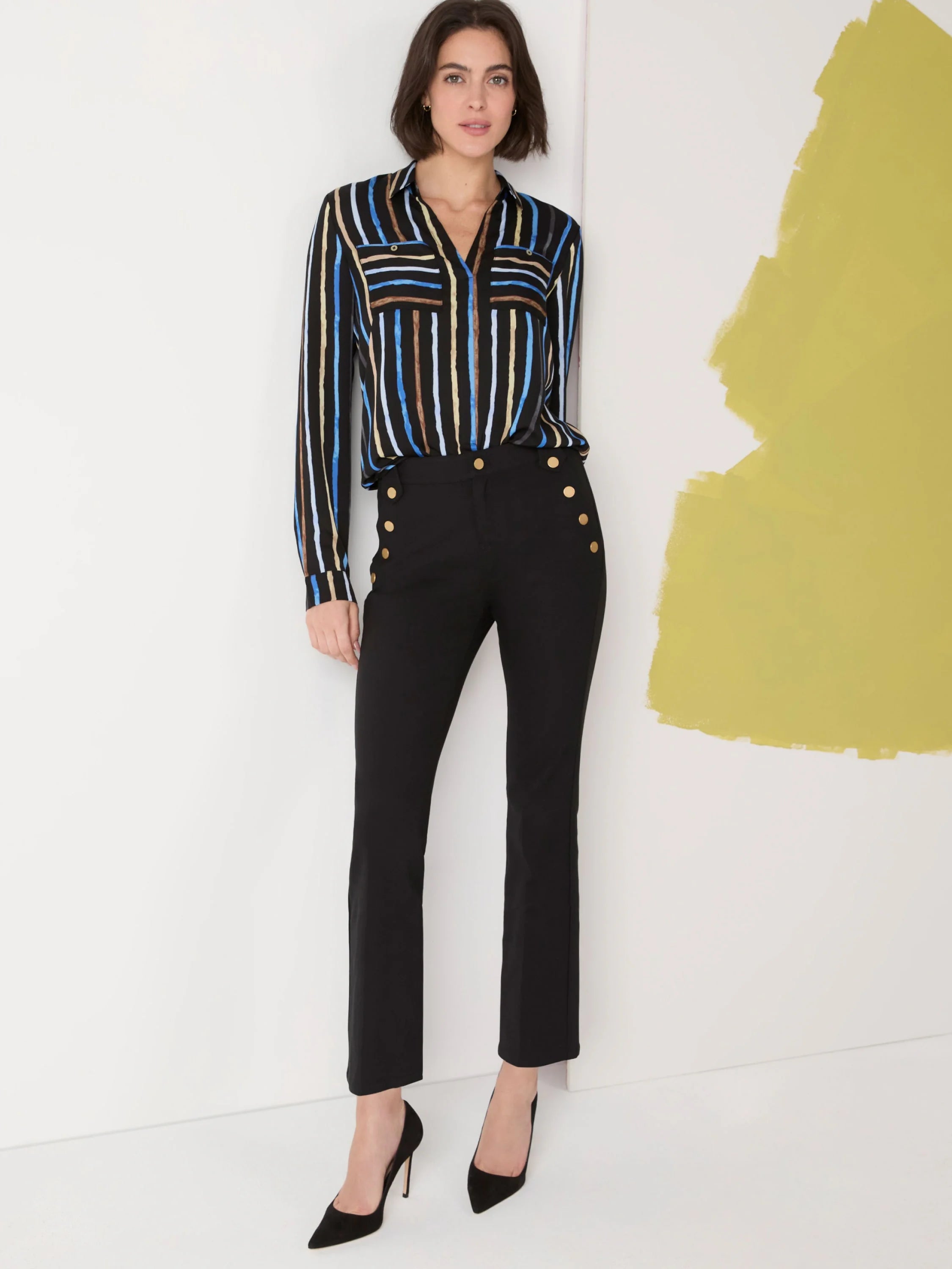 Black mid-rise pants, slightly flared at the ankle, with gold buttons. Model also wears a black and blue striped shirt.