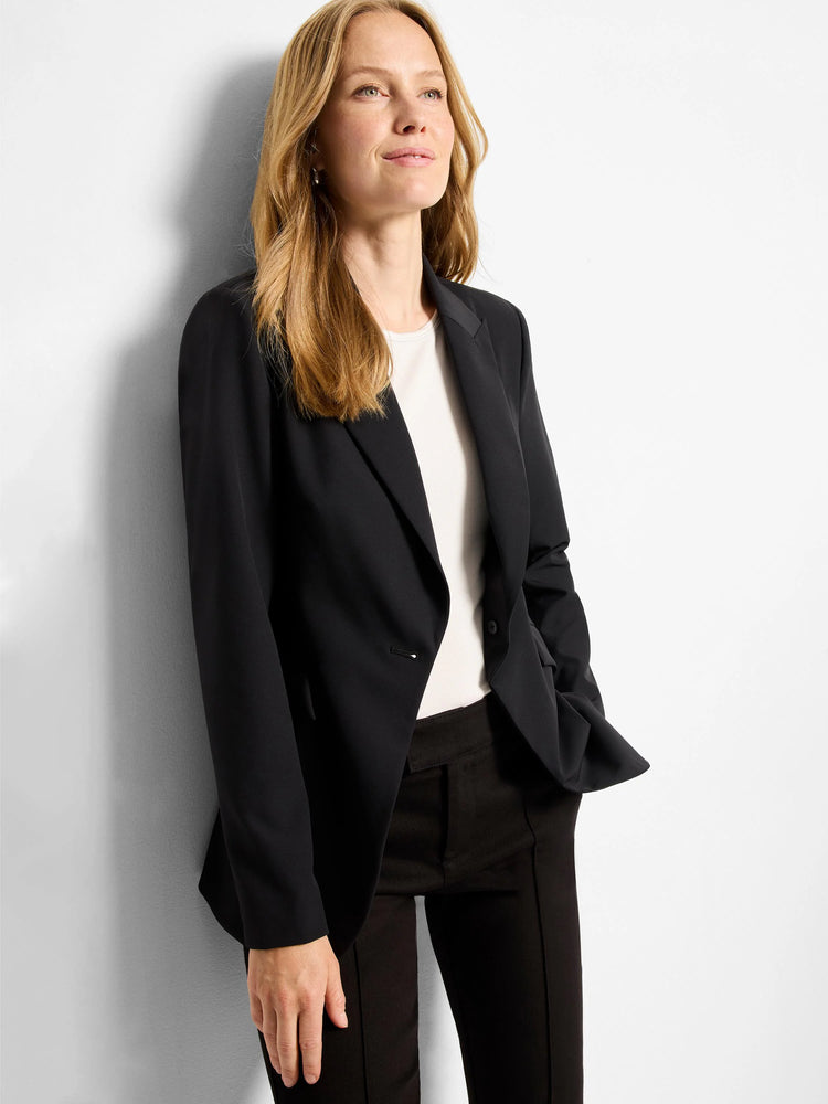 Black single-breasted blazer; notched lapel. Single button closure. Model wears it with dark trousers.