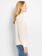 Creamy white chiffon long-sleeve top with a draped neckline and button cuffs, worn by a model with blue jeans.