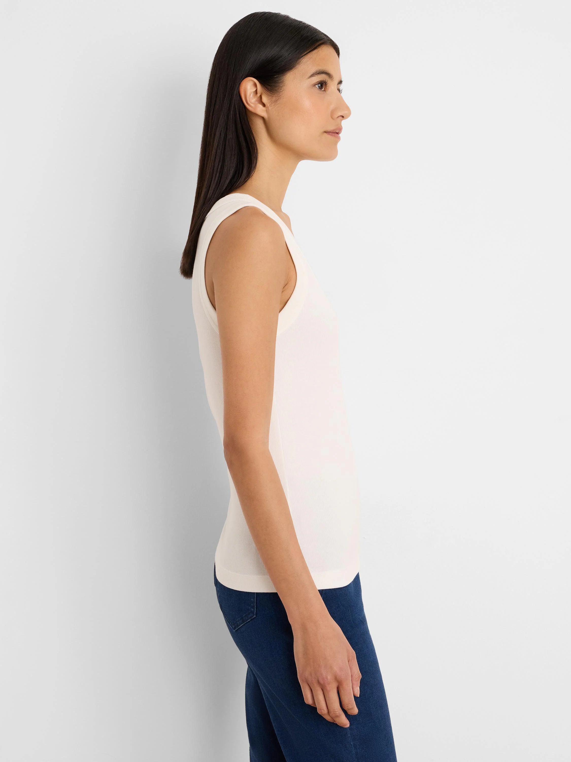 Off-white ribbed scoop neck tank top, worn by a model in profile. Dark blue jeans are visible.