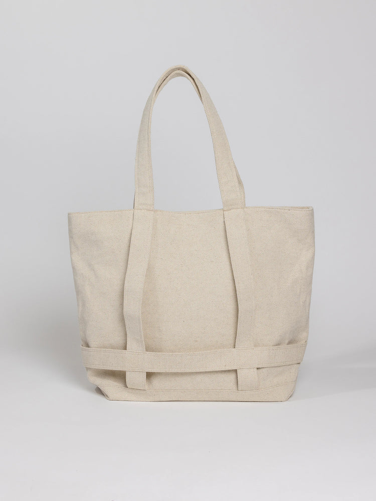 Off-white canvas tote bag with two top handles and a reinforced bottom. The bag features a woven detail across the bottom.