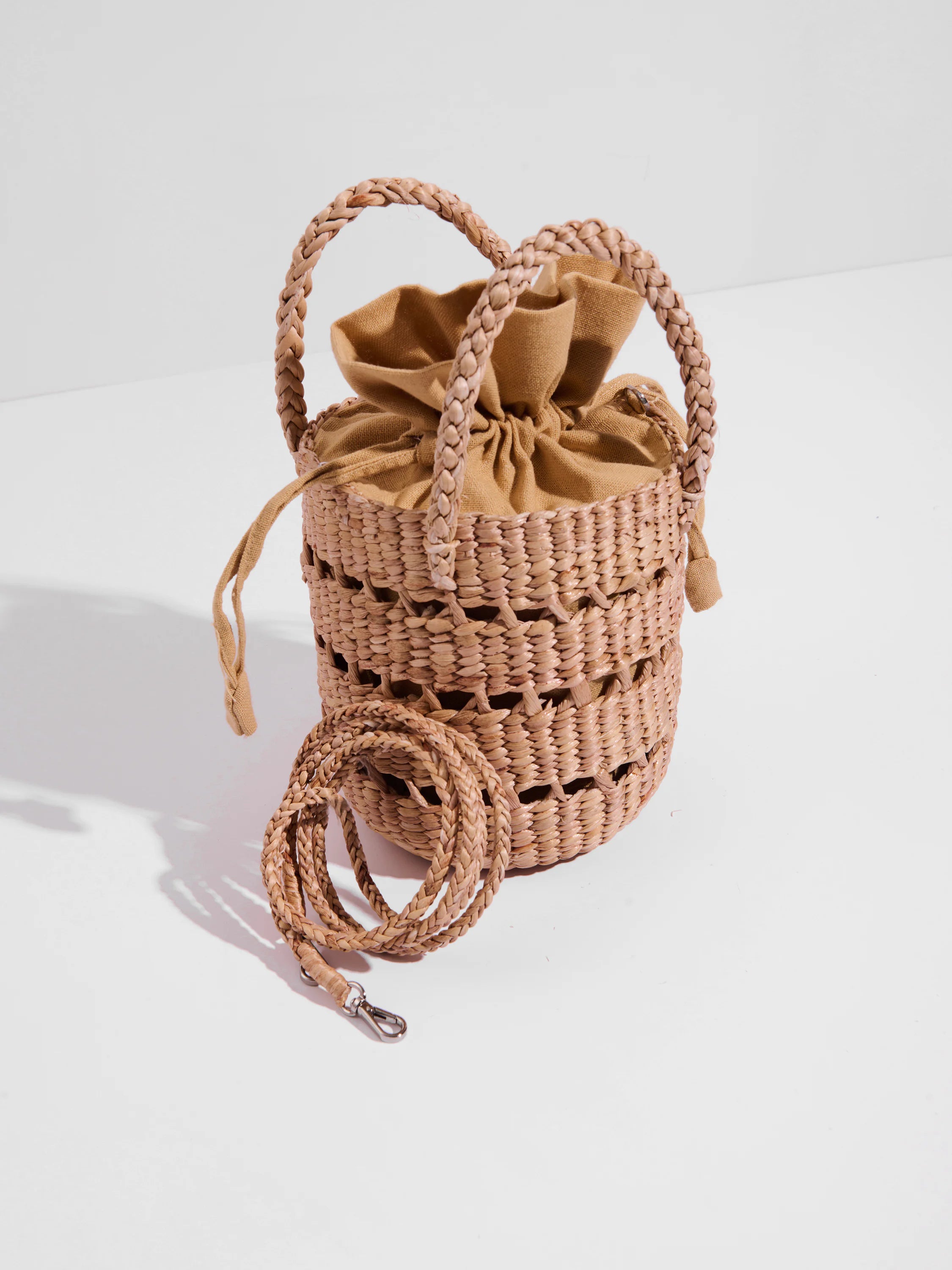 Light brown woven cylindrical bag with braided handles, light tan drawstring top, and a matching braided crossbody strap.