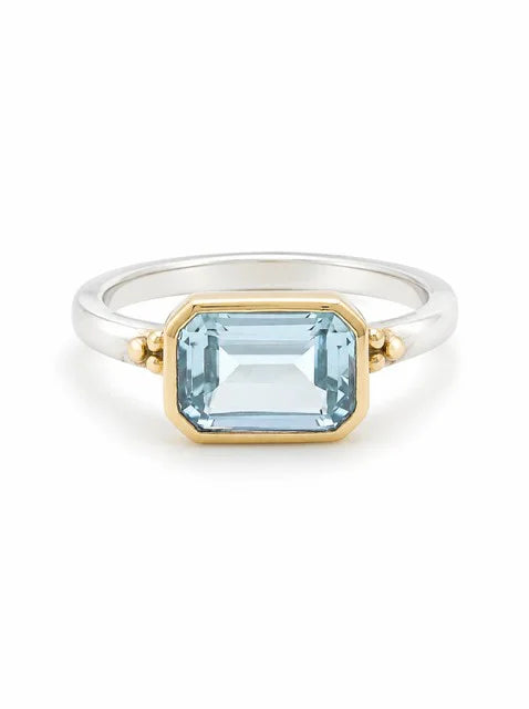 Silver ring with gold bezel-set emerald-cut light blue stone, flanked by small gold beads.