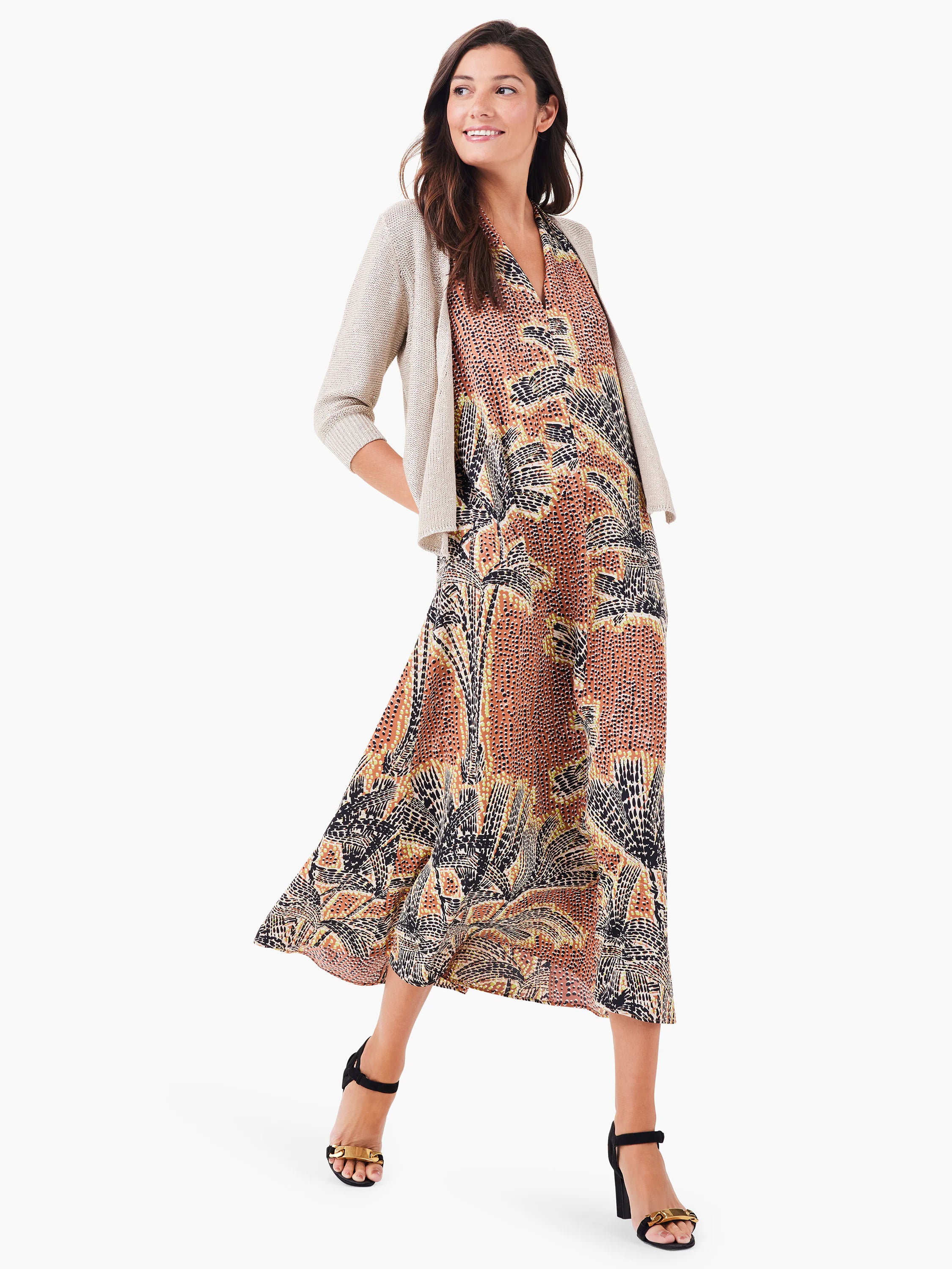 Maxi dress with brown, black, and beige palm tree print. Halter neck, A-line silhouette. Model wears a beige cardigan.