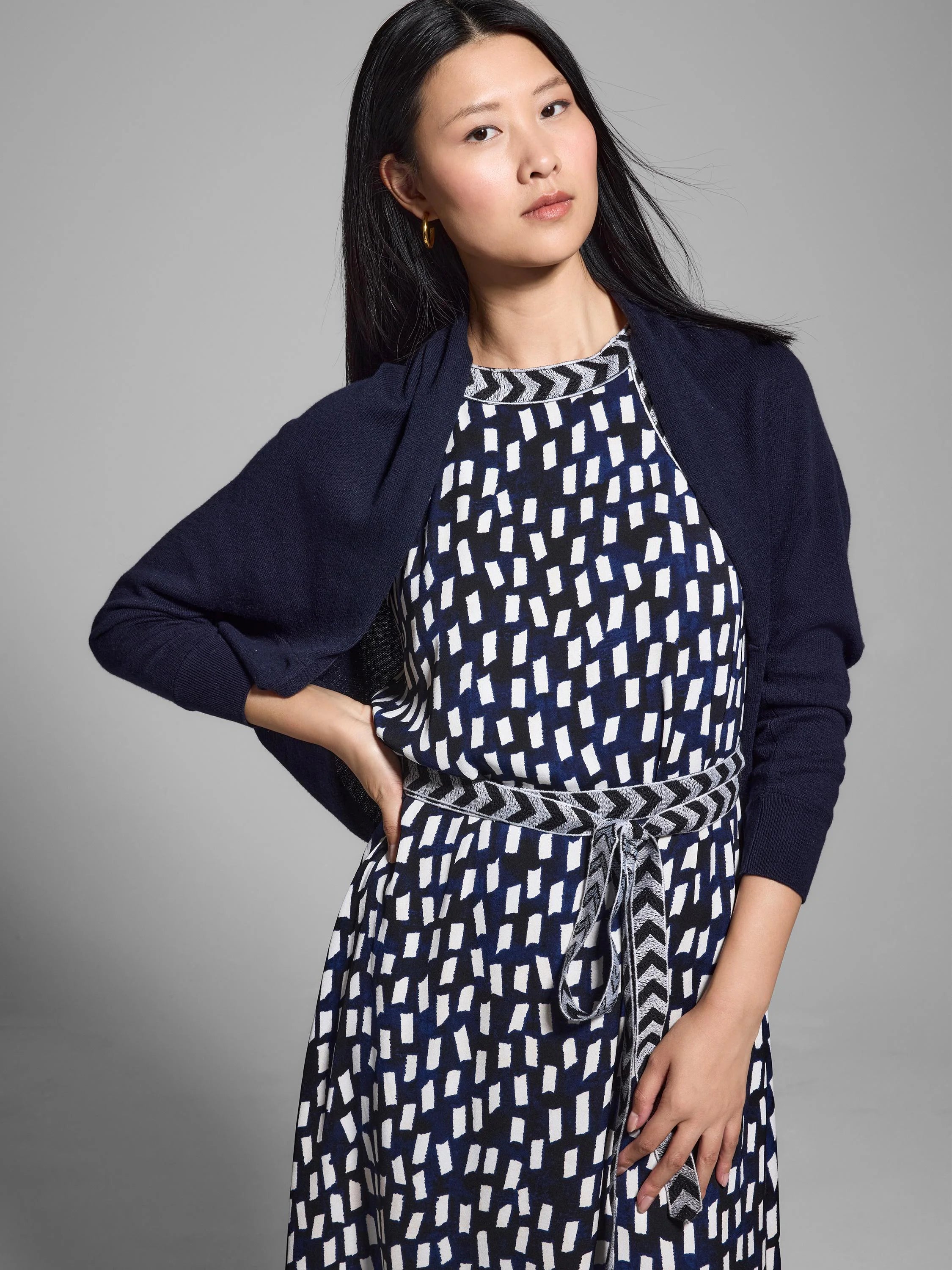 Navy blue shrug cardigan; three-quarter sleeves. Worn over white and navy dress with geometric pattern and gray belt.