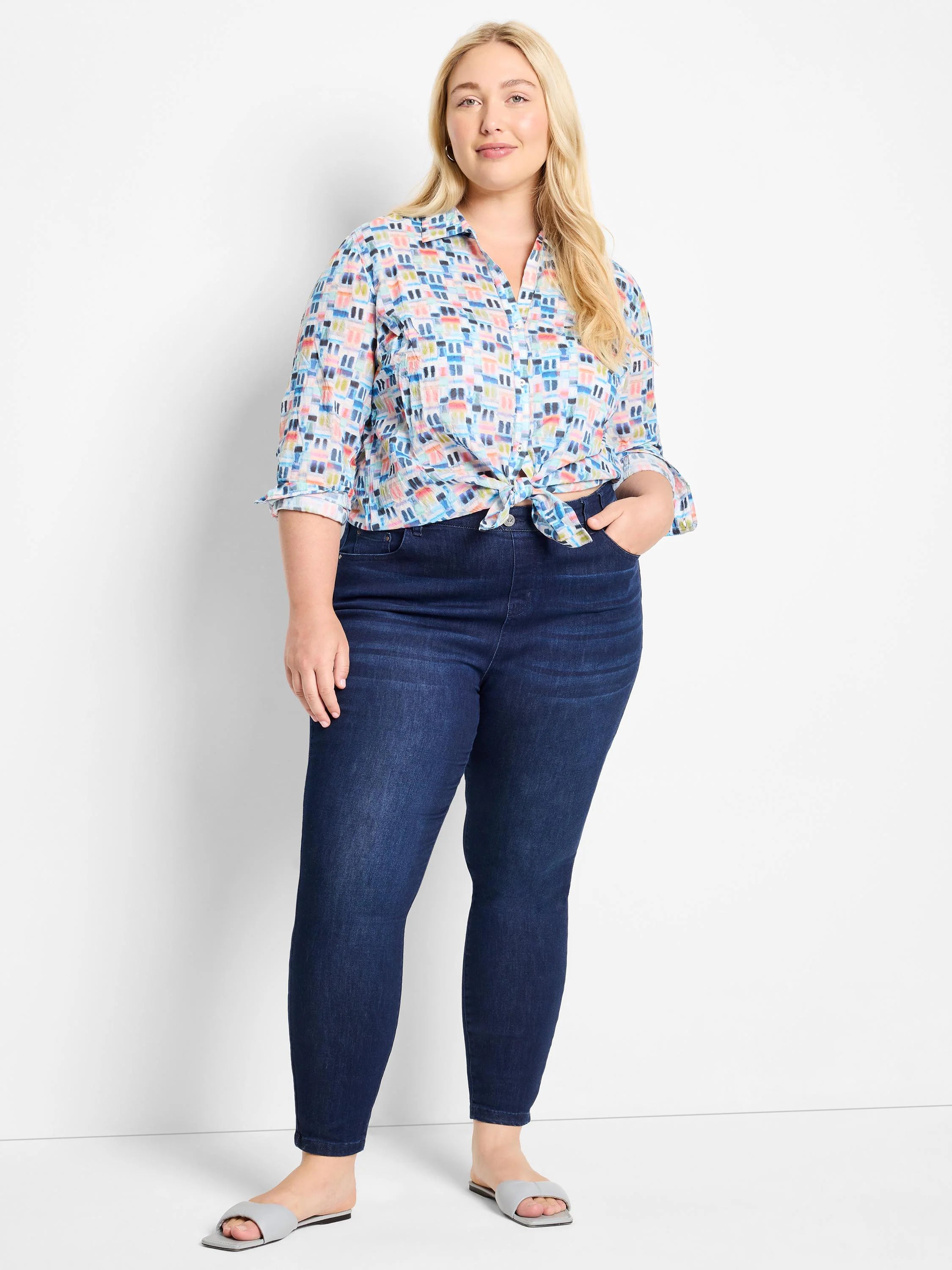 Woman wearing a light blue, multicolored-geometric print boyfriend shirt knotted at the waist, dark wash jeans, and light...