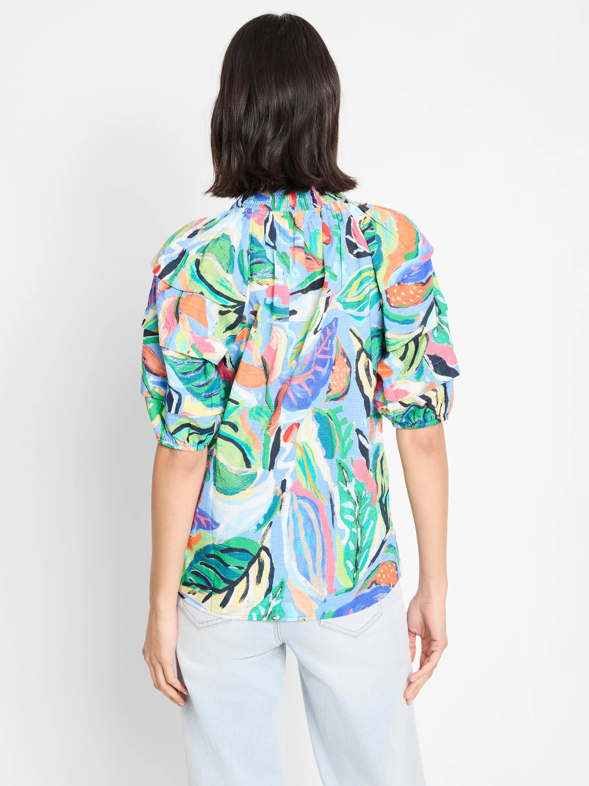 Light blue top with short sleeves and ruffle details. Multicolored abstract leaf print in greens, pinks, oranges.
