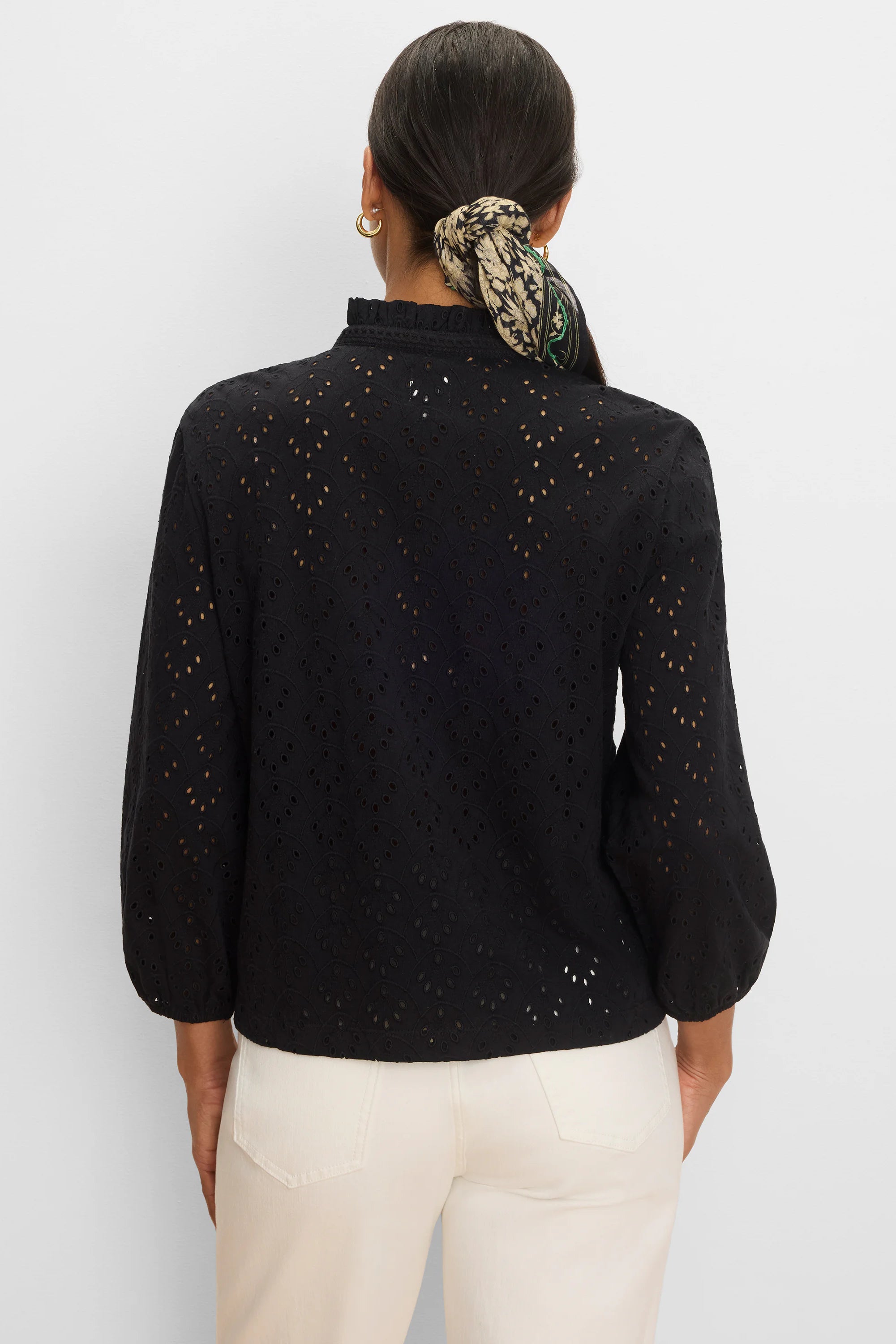 Black knit top with a ruffled high neck, long voluminous sleeves, and an all-over scalloped eyelet pattern.
