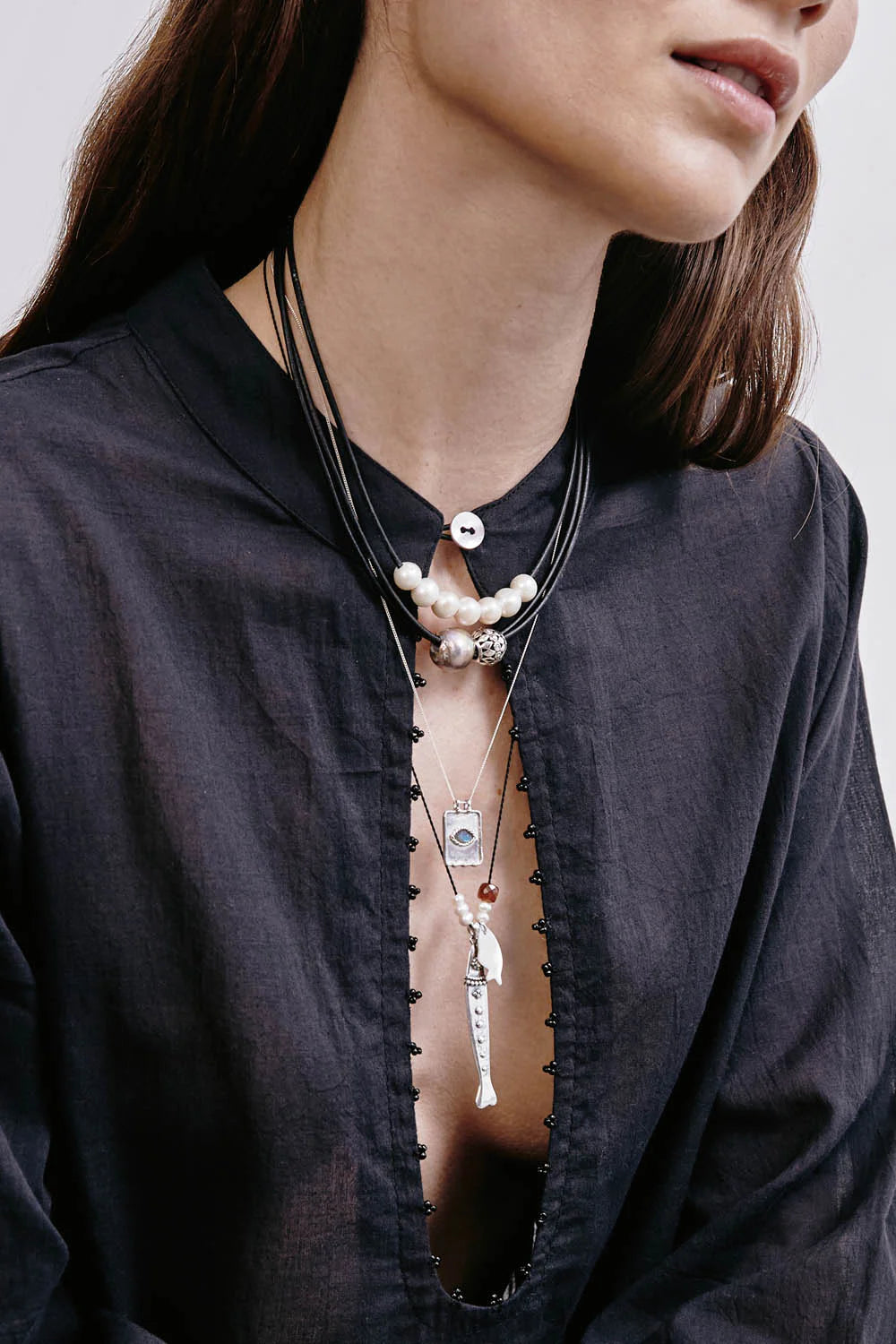 Black cord necklace with silver fish charm, white pearl, and reddish bead. Worn with other layered necklaces.