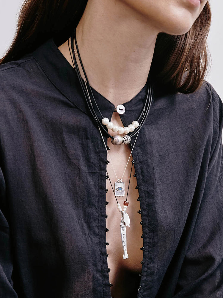 Silver fish charm necklace on black cord, layered with pearl and silver bead necklaces. Features small silver...
