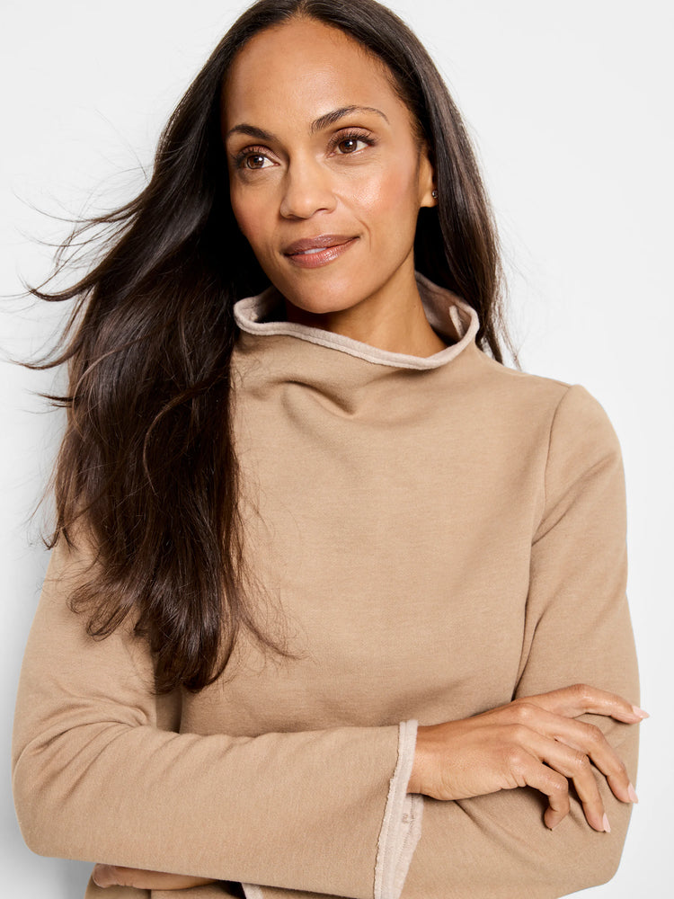 Beige long-sleeve top with lighter beige trim at the neck and cuffs. Mock neck. Model is wearing the top with arms crossed.