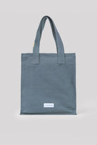 Slate blue cotton tote bag with two fabric handles and a white "LES PENSIONNAIRES" label on the front.
