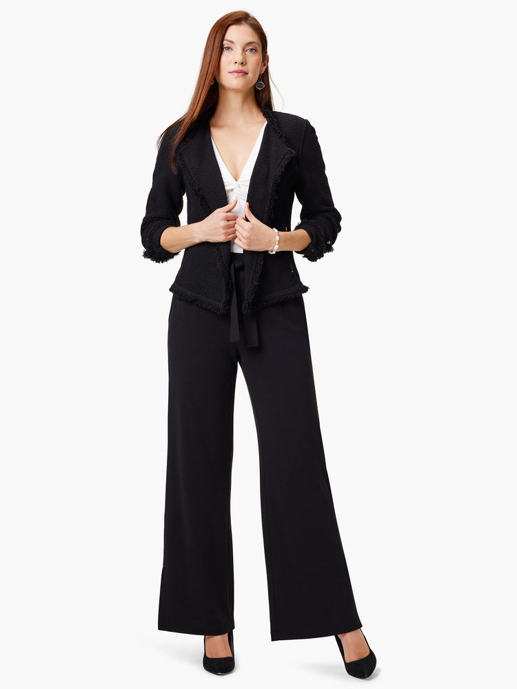 Black textured jacket with fringed edges over a white top. Model also wears black pants and shoes.