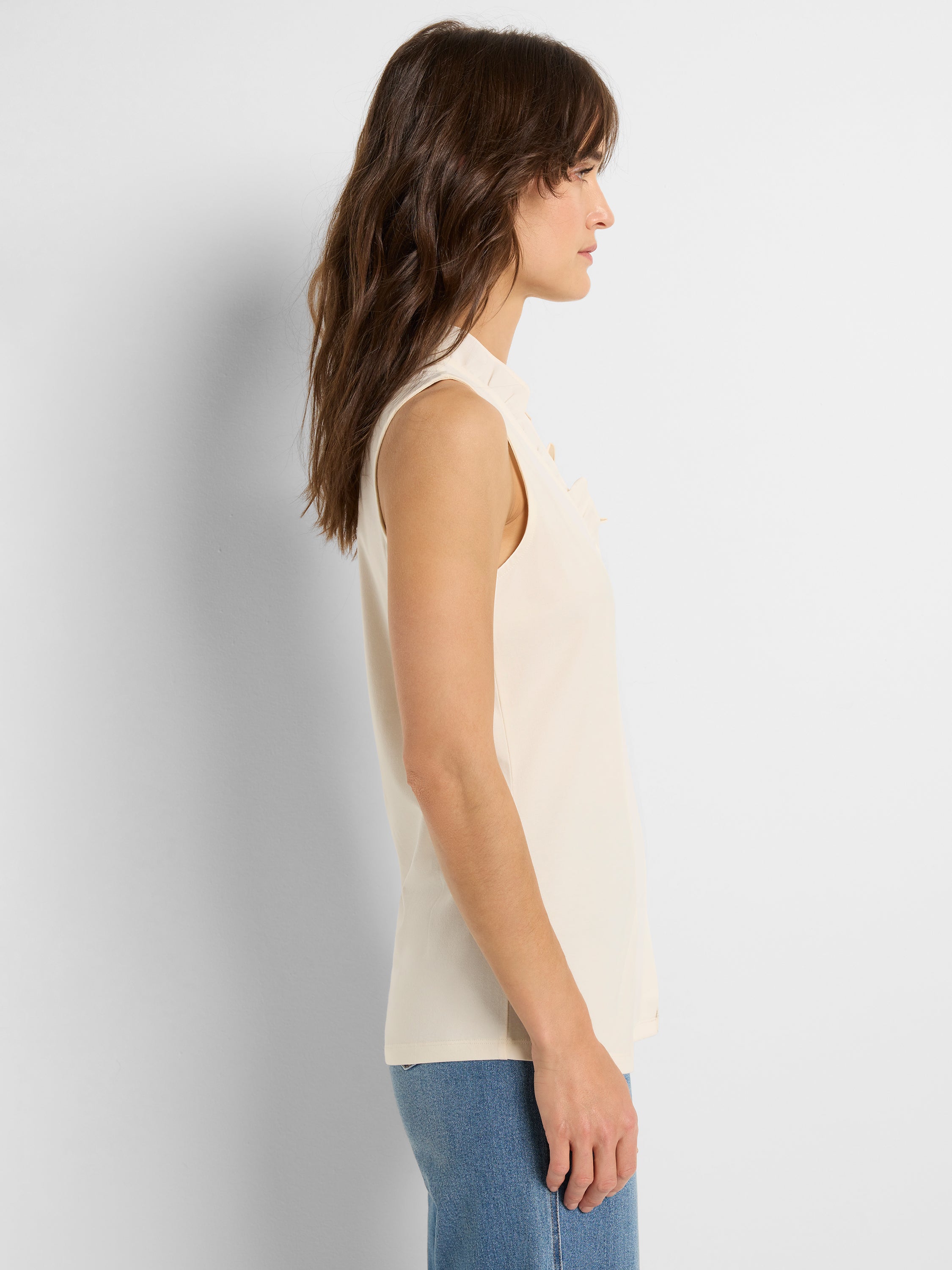 Cream sleeveless blouse featuring a straight fit and a small standing collar with a tie detail, styled with denim jeans.