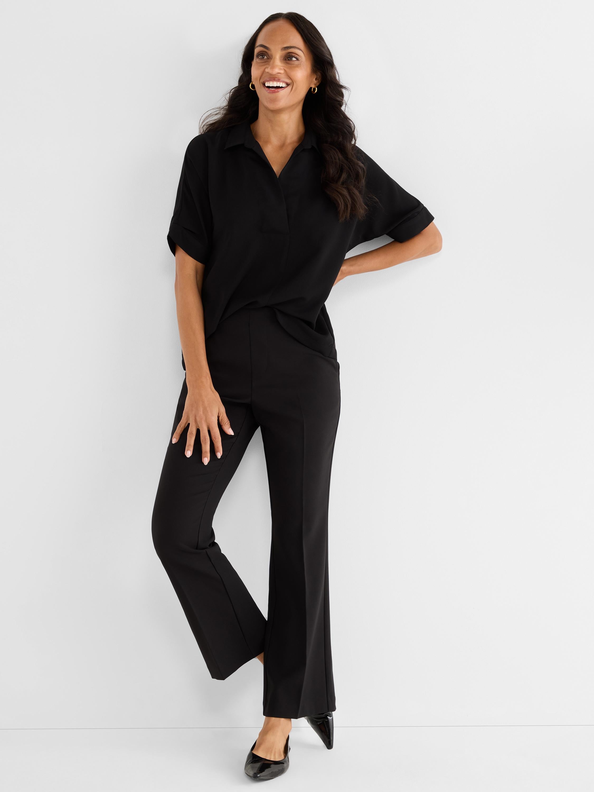 She wears a relaxed black short-sleeve collared top paired with high-waisted tailored black bootcut pants.