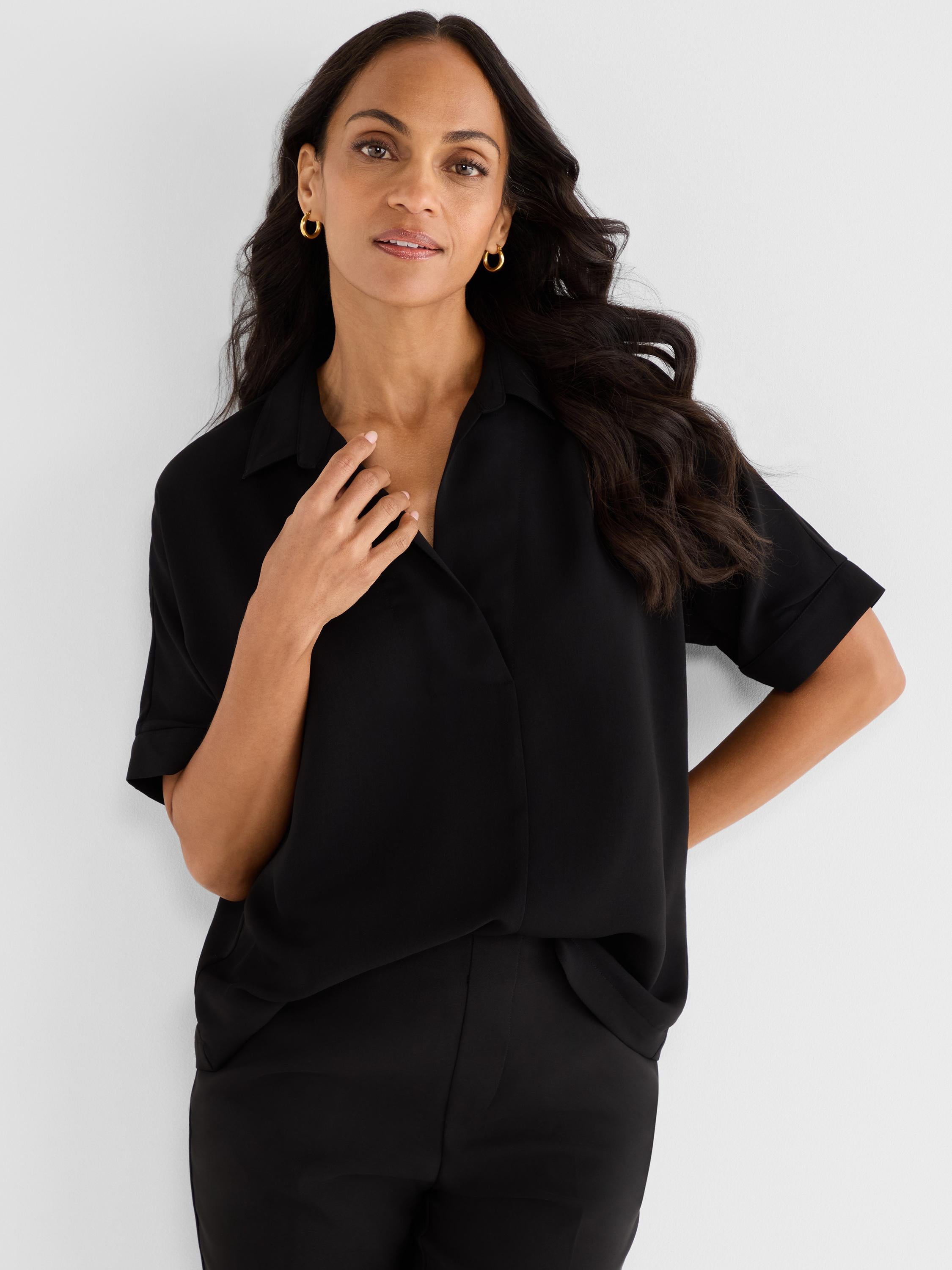 A black short-sleeve draped blouse with a relaxed fit and open collar is styled professionally with black trousers.