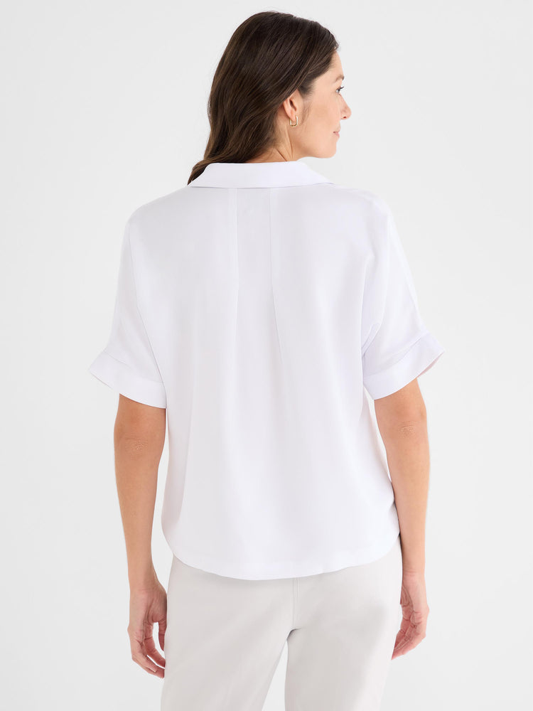 Model wears a relaxed white collared blouse with cuffed short sleeves and a structured back seam.