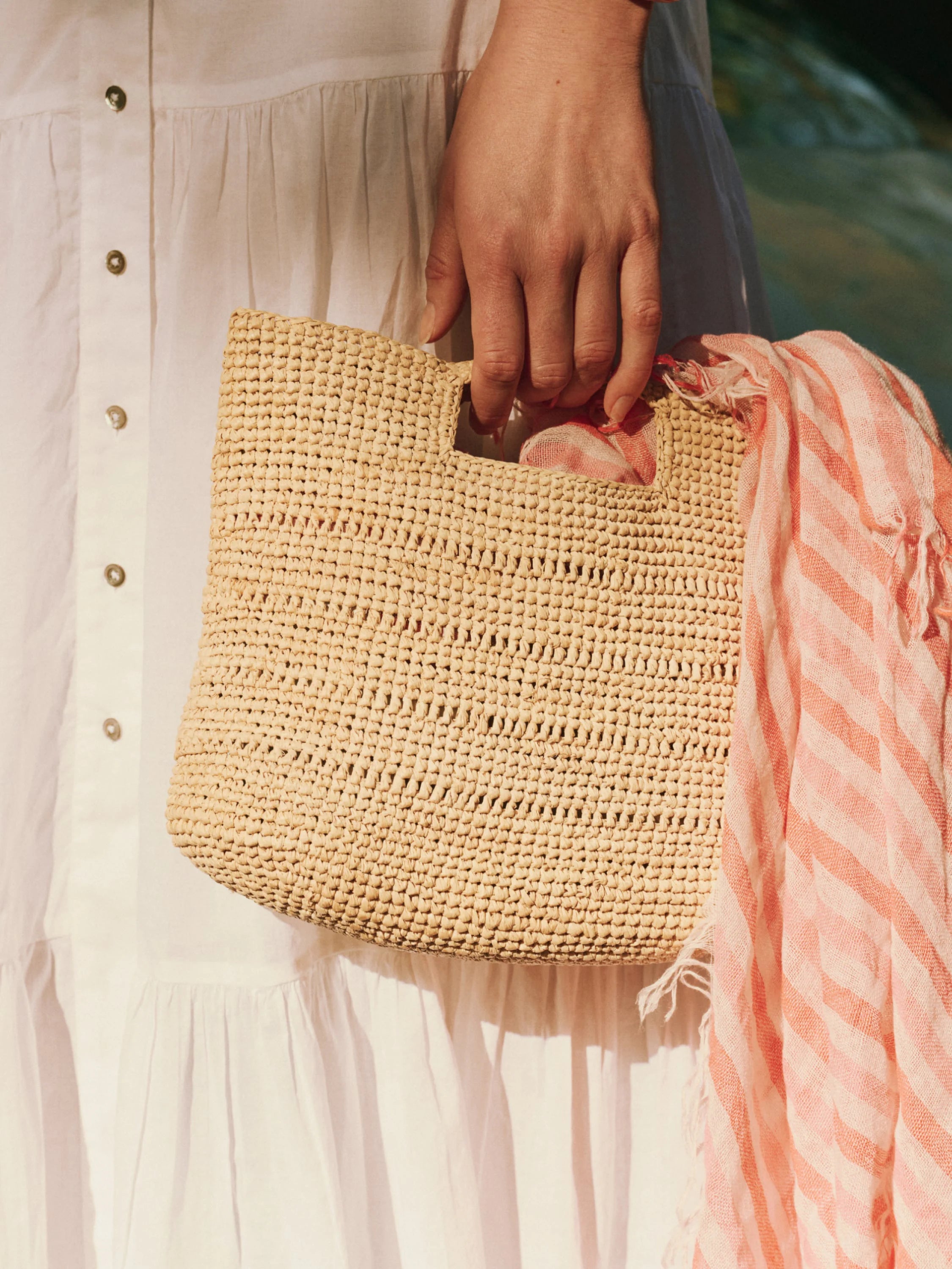 Natural woven straw rectangular handheld bag with cut-out handles, held by hand. A pink and white striped scarf is inside.