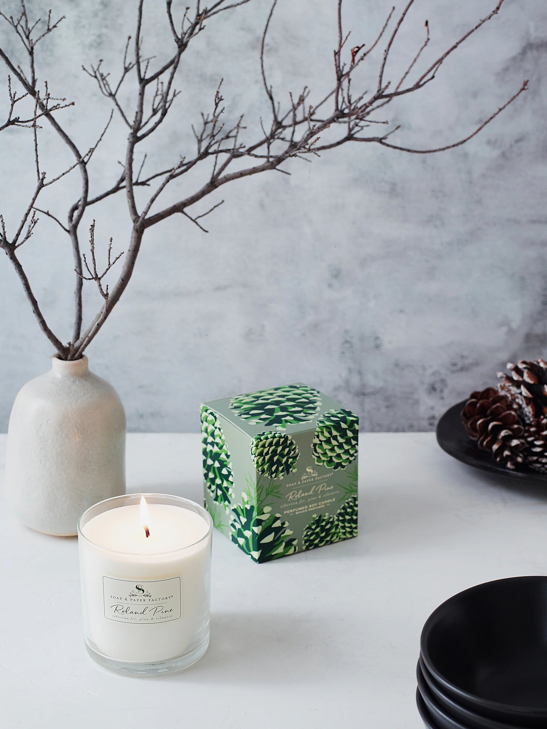 White candle in clear glass, lit, next to its sage green box featuring a pinecone pattern.
