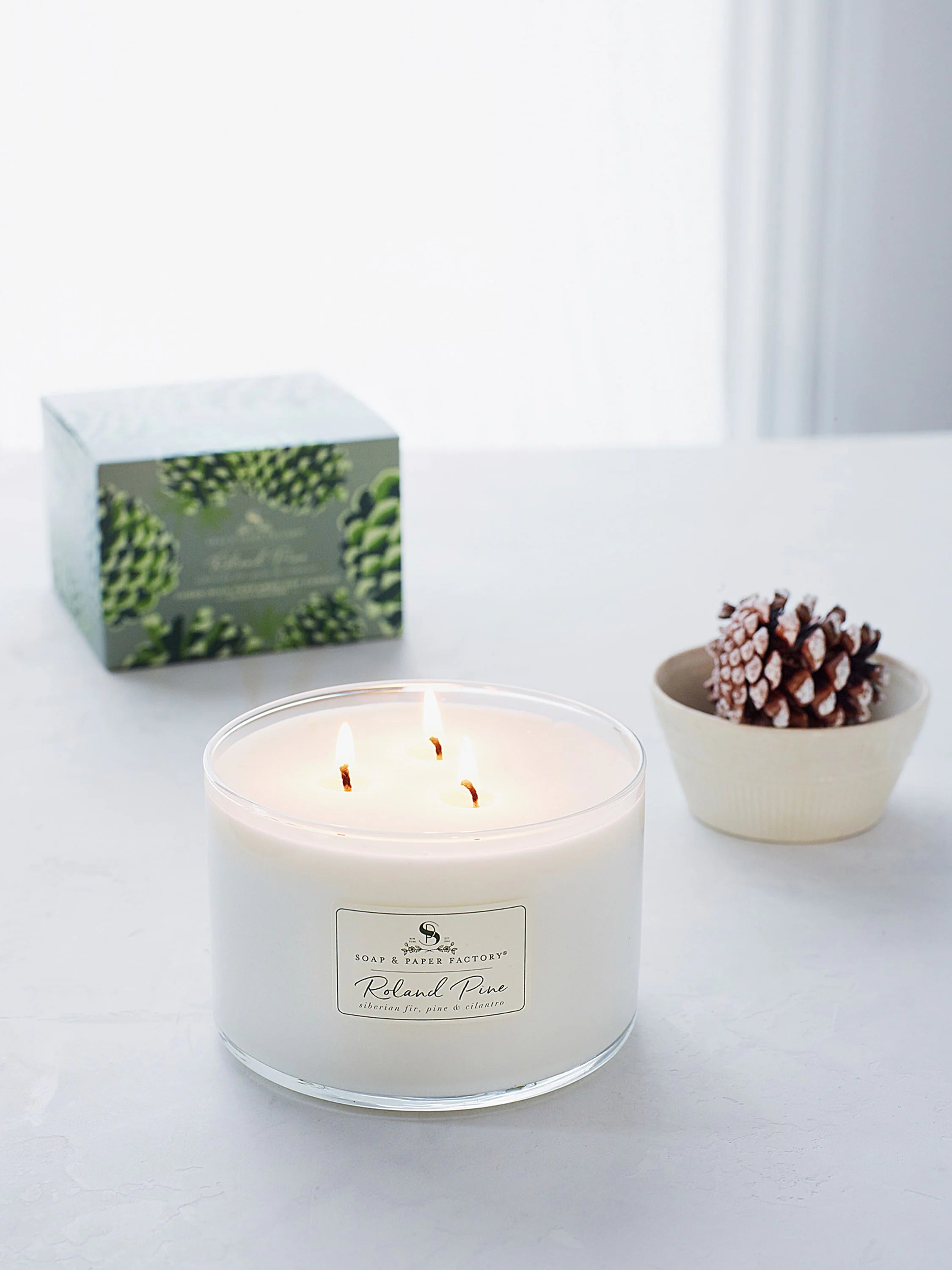 White soy candle in clear glass, three lit wicks. Label shows green pine cone design and product name.