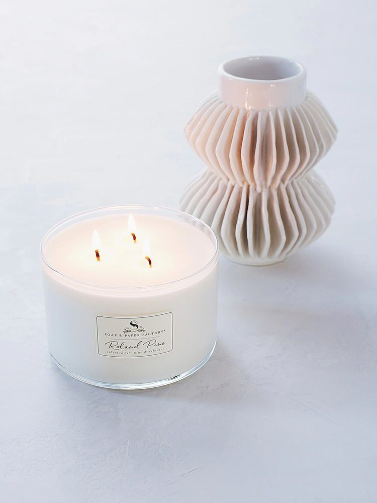 Off-white soy candle in clear glass, three lit wicks. Label shows product name. Next to a white pleated vase.
