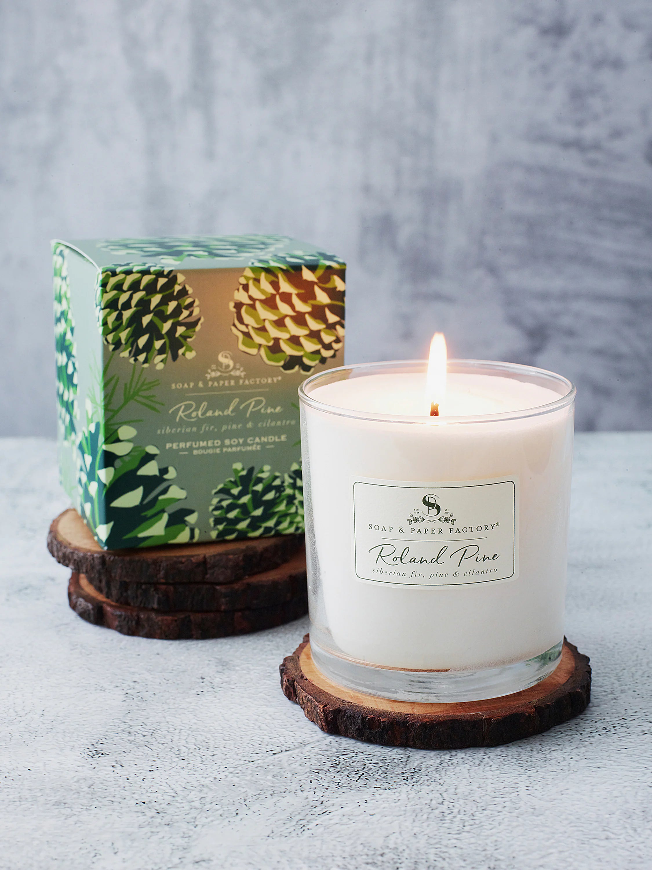 White soy candle in clear glass, burning, on wood slices. Box shows green & beige pine cone design.