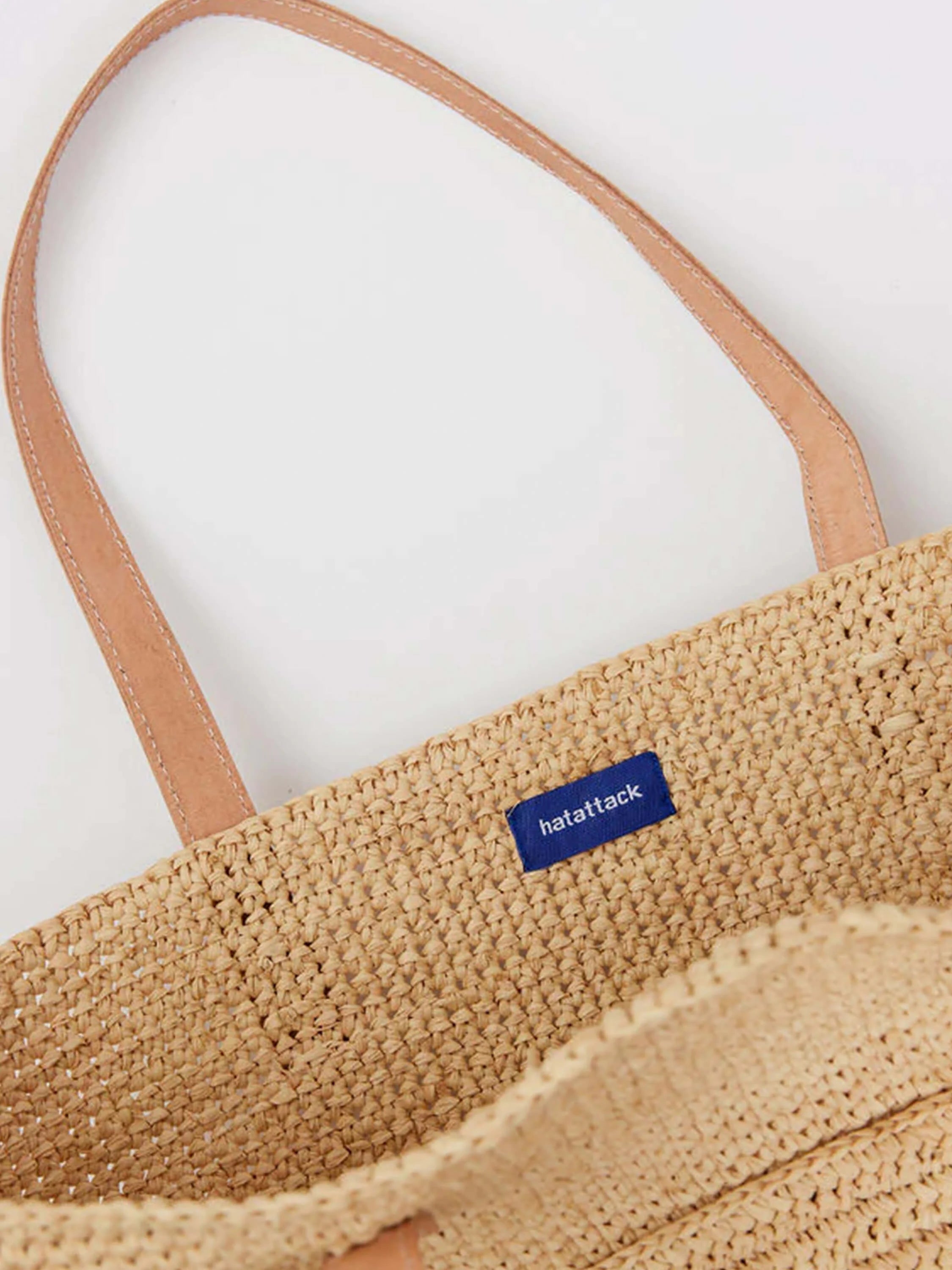 Tan woven raffia tote, light brown leather handles with white stitching, blue 'hatattack' tag.