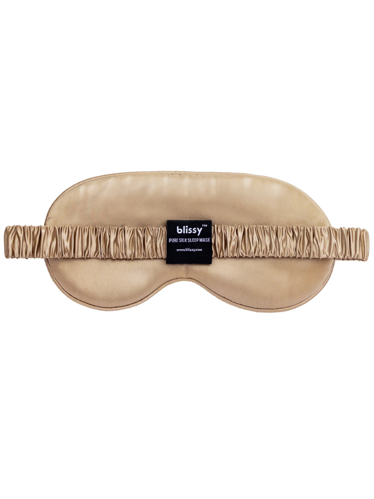 Champagne-colored contoured silk sleep mask with a ruched elastic strap and black brand tag.