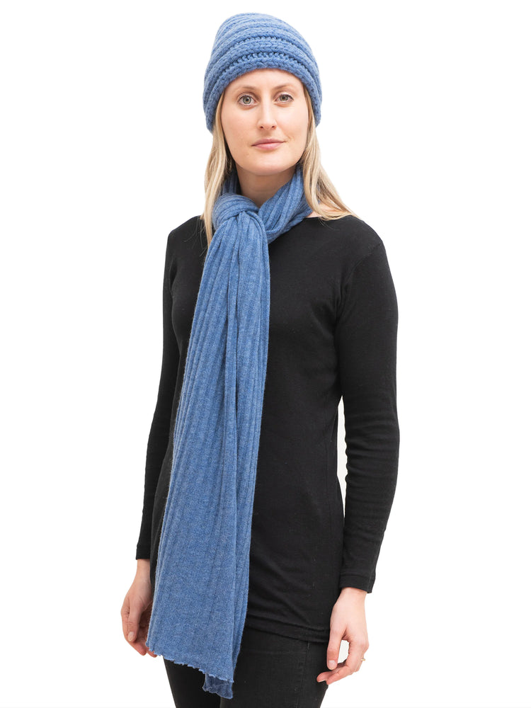 Blue cashmere scarf with ribbed texture, shown loosely draped. Model wears black top and matching blue hat.