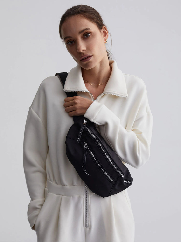 Black belt bag with three silver front zippers, white 'VARLEY' logo, and black pulls with white stitching, worn across torso.