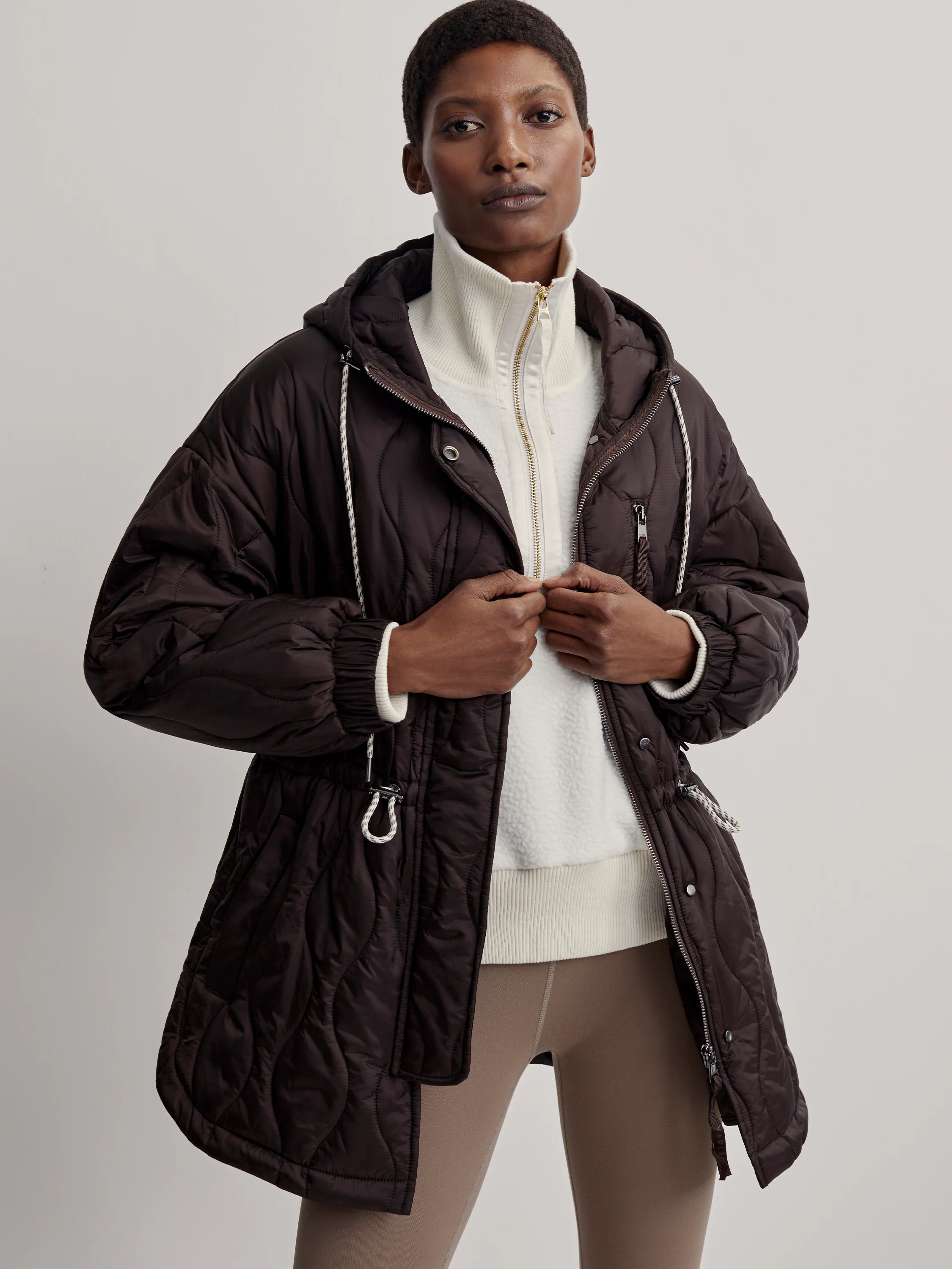 Dark brown hooded quilted jacket features wavy stitching, white drawstrings, and front zipper closure.