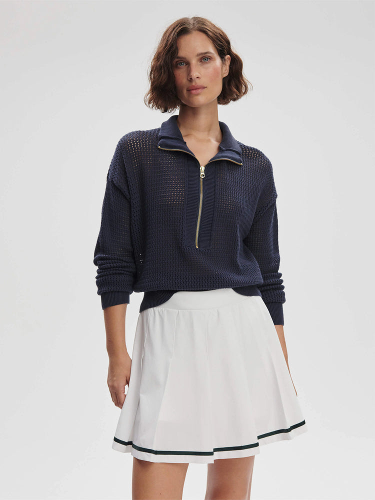 Navy blue, half-zip, open knit sweater; gold zipper. Model also wears white pleated skirt with dark green trim.