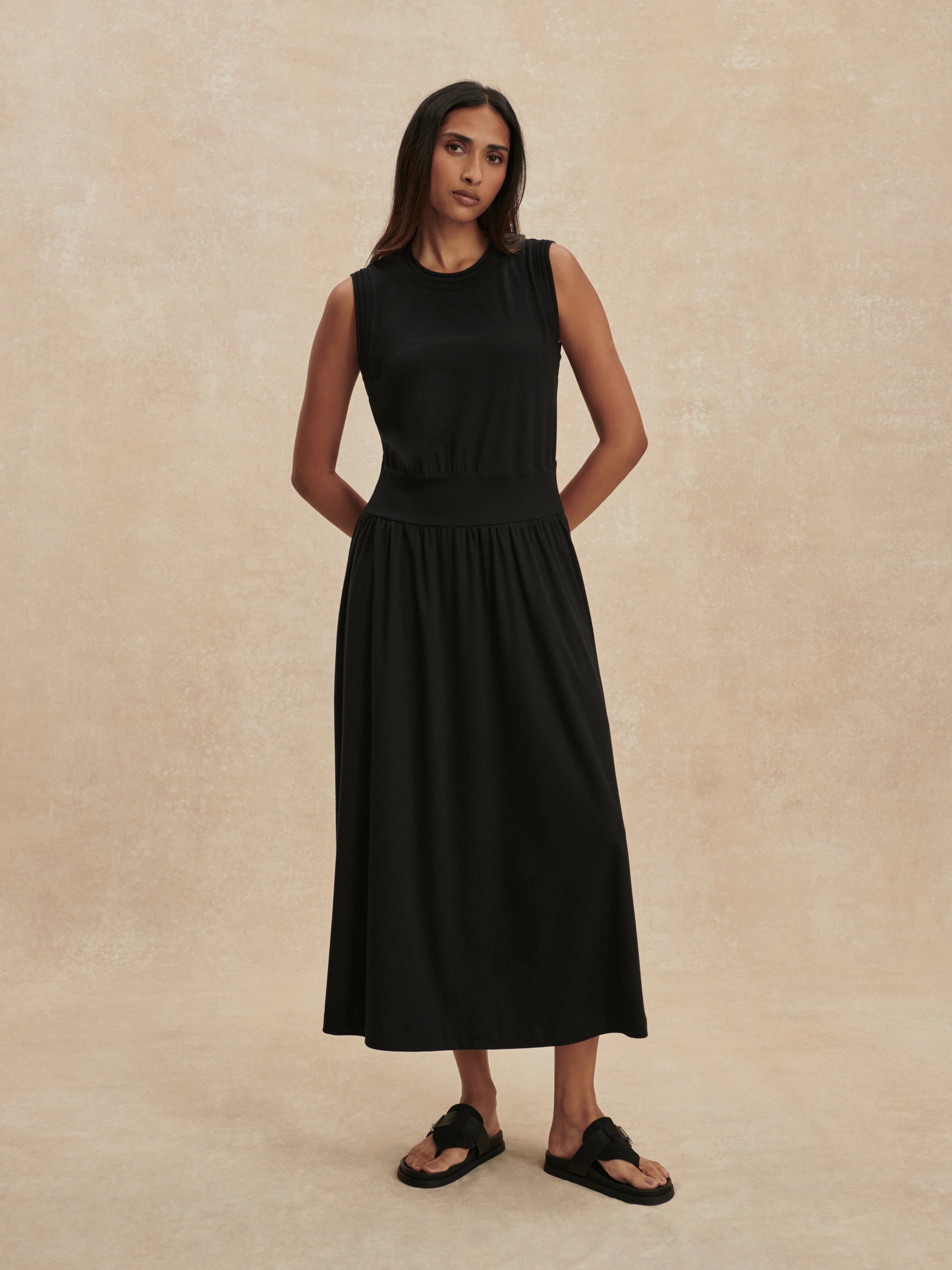 Black sleeveless maxi dress with round neck, fitted bodice, elasticated waist, and a long, flowing skirt.